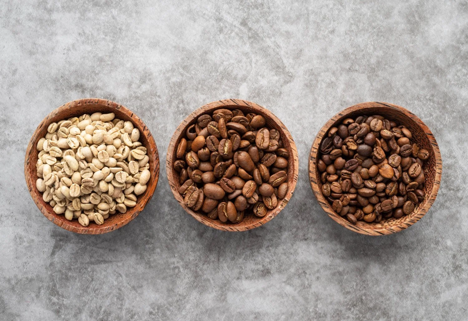 Three small wooden bowls filled with different types of coffee beans arranged in a row on a gray surface.