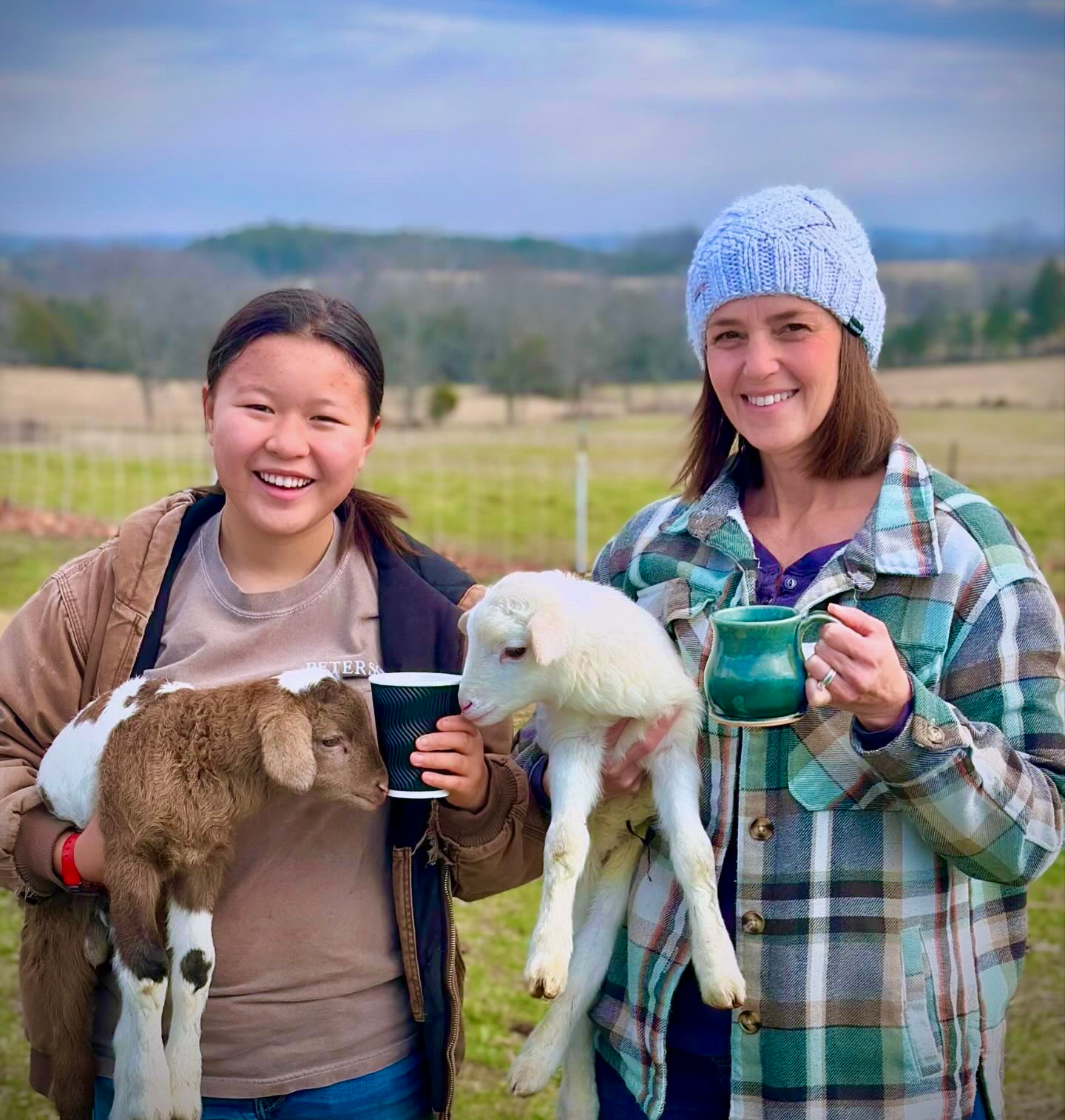 Two women standing outside on a farm holding baby goats with a rural landscape in the background. One woman is holding a brown and white goat, and the other is holding a white goat. Both women are smiling and holding cups.