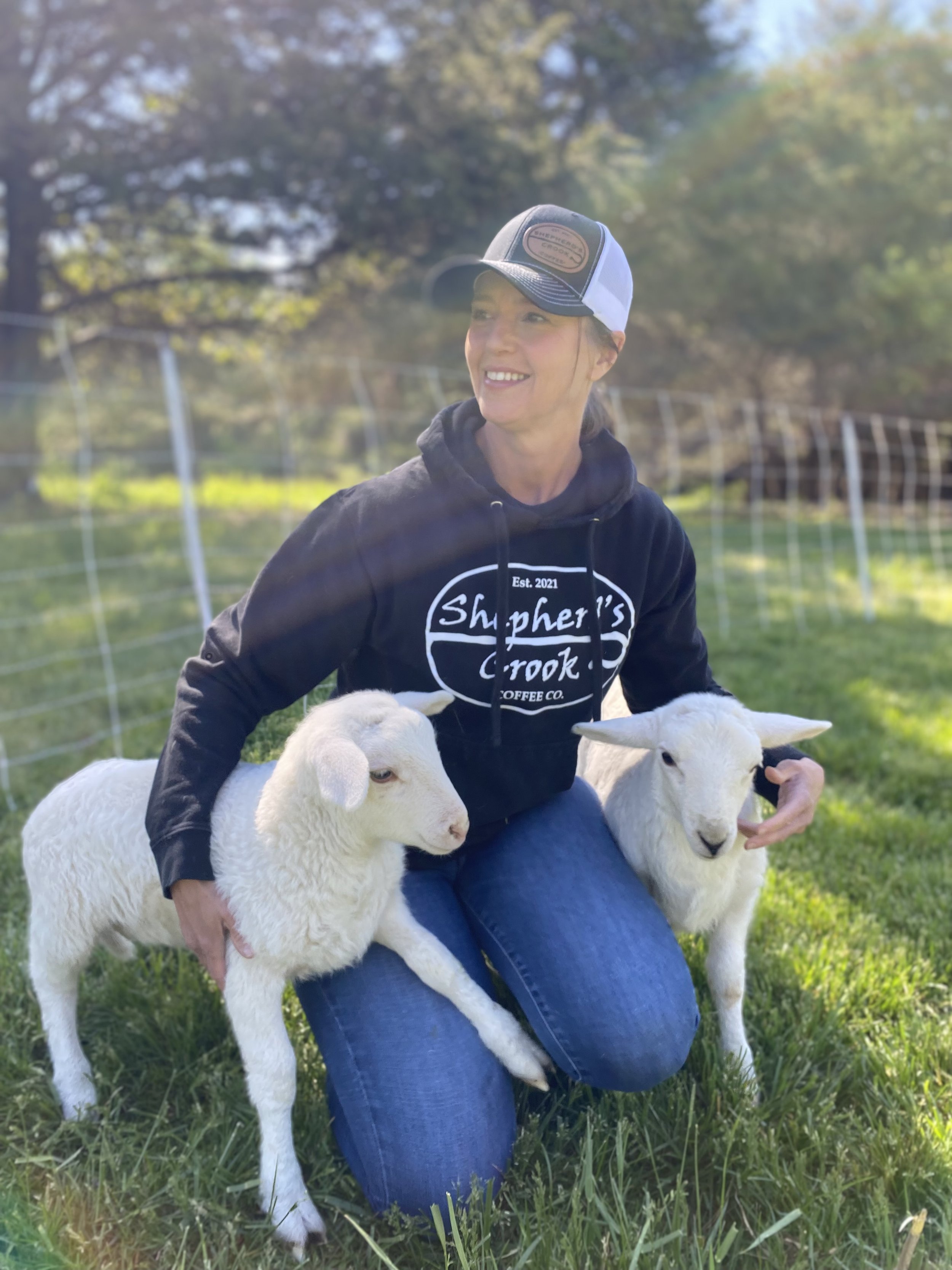 A woman kneeling on grass, holding two young white goats, with a fence and trees in the background.