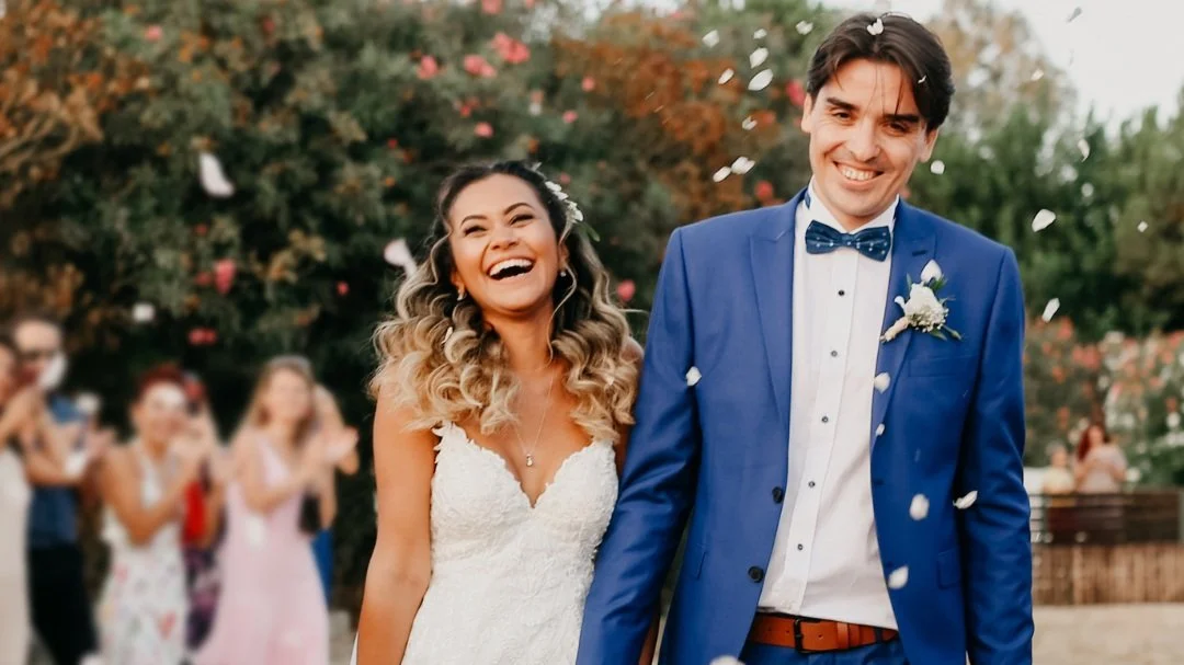 Happy bride and groom smiling after wedding ceremony, with guests celebrating in background and flower petals falling.