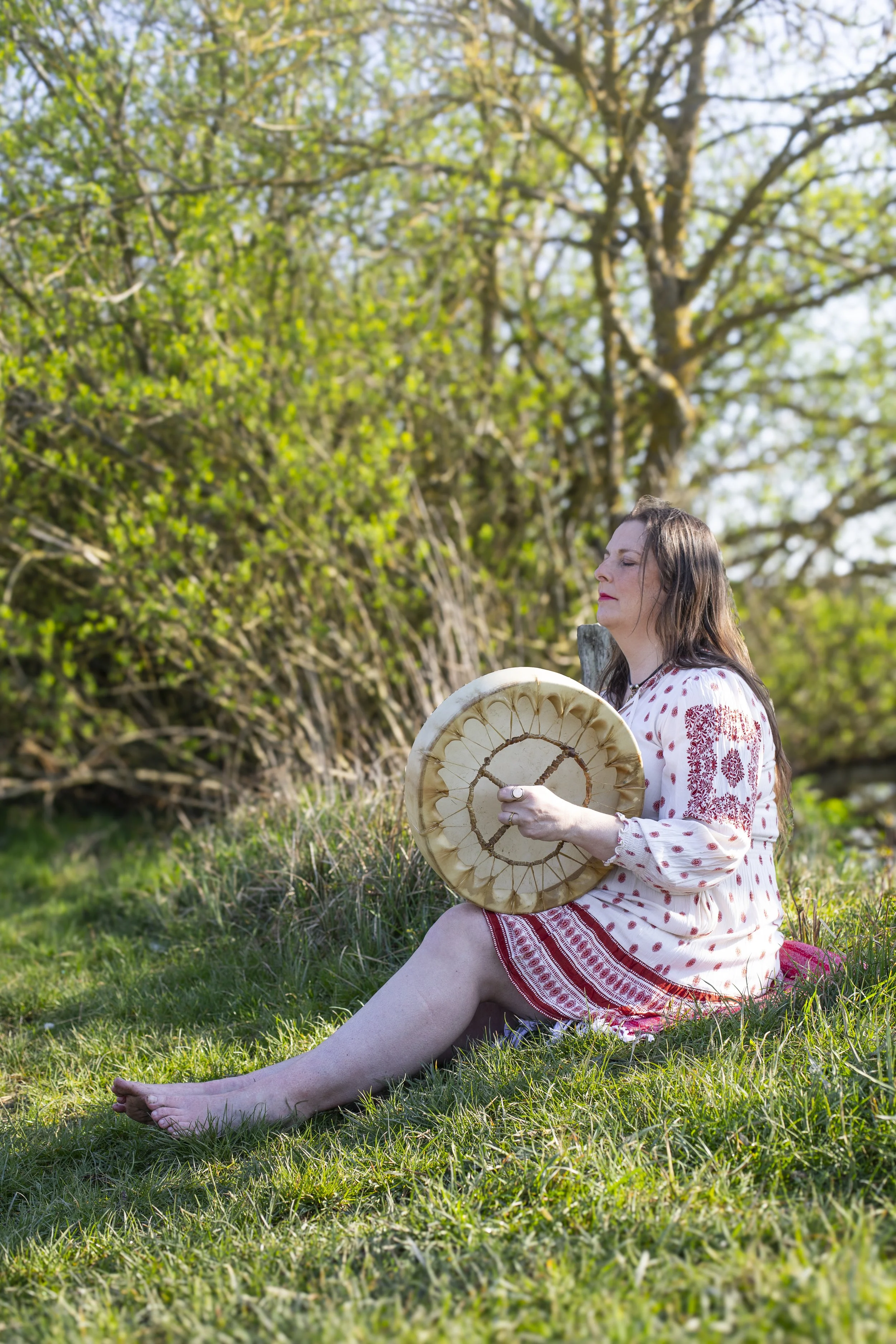 A woman with closed eyes sitting outdoors on grass holding a large circular object that resembles a piece of tree trunk or a carved wooden disk. She is wearing a white dress with red embroidery and has long brown hair. In the background, there is a lush green tree with small leaves and a partly cloudy sky.