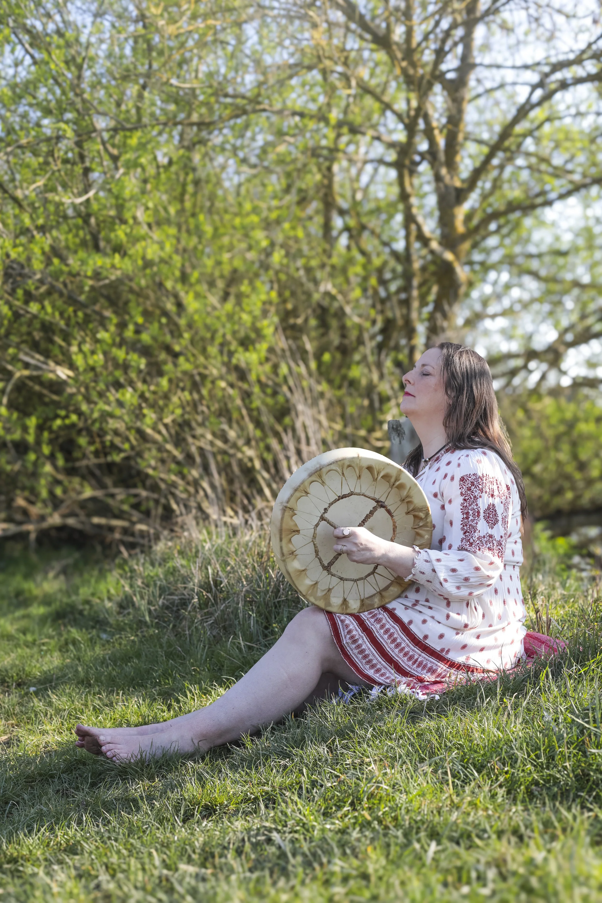 A woman in a white and red embroidered dress sits on the grass outdoors, holding a large, round, cream-colored drum. She appears relaxed and peaceful with her eyes closed and face tilted slightly upward. Behind her, there is a lush green tree with bright leaves and sunlight filtering through.