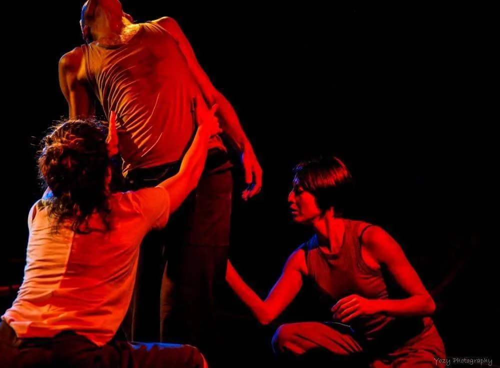 Three women are helping a man onto a stage or platform; the scene is illuminated with red and yellow lighting, and the background is dark.