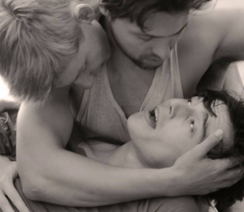 Three young men are engaged in playful, close contact, with two of them leaning in towards the third, who is lying down and looking up at them, smiling.