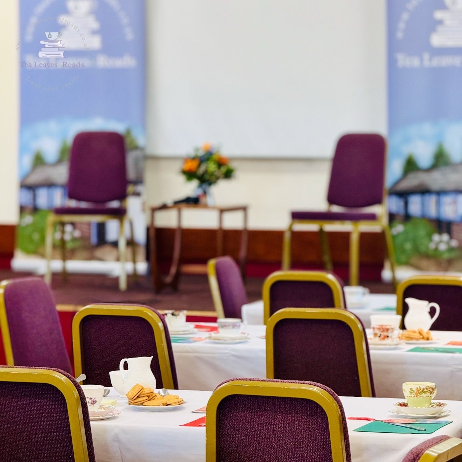Conference room with round tables set with tea cups, plates of cookies, and placemats. Chairs with purple upholstery and yellow frames surround the tables. In the background, there are two purple chairs, a small table with a flower arrangement, and a whiteboard. Colorful banners or posters are on the walls.