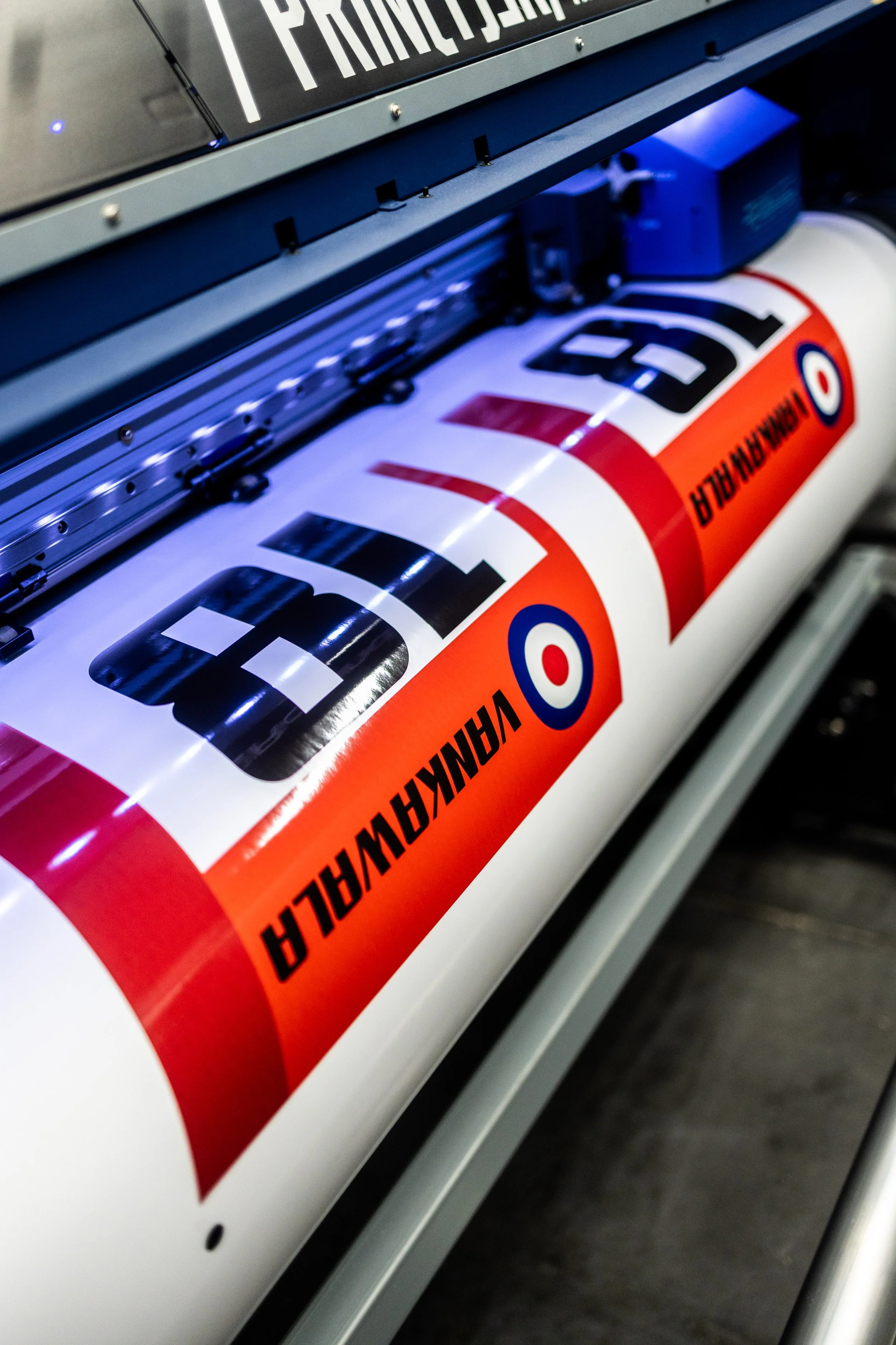 A large printing machine producing a race car livery with the word "F1" and the color scheme of orange, white, black, and blue roundel, related to Formula 1 racing.