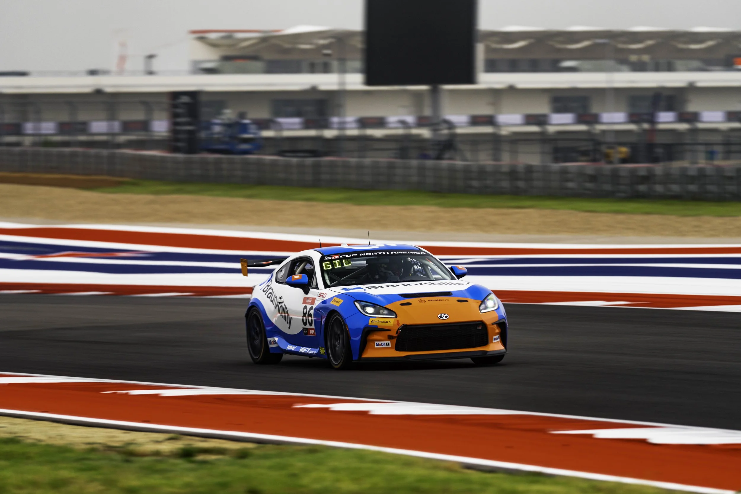A racing car on a track during a race, with a blurred background of the stands and sky.