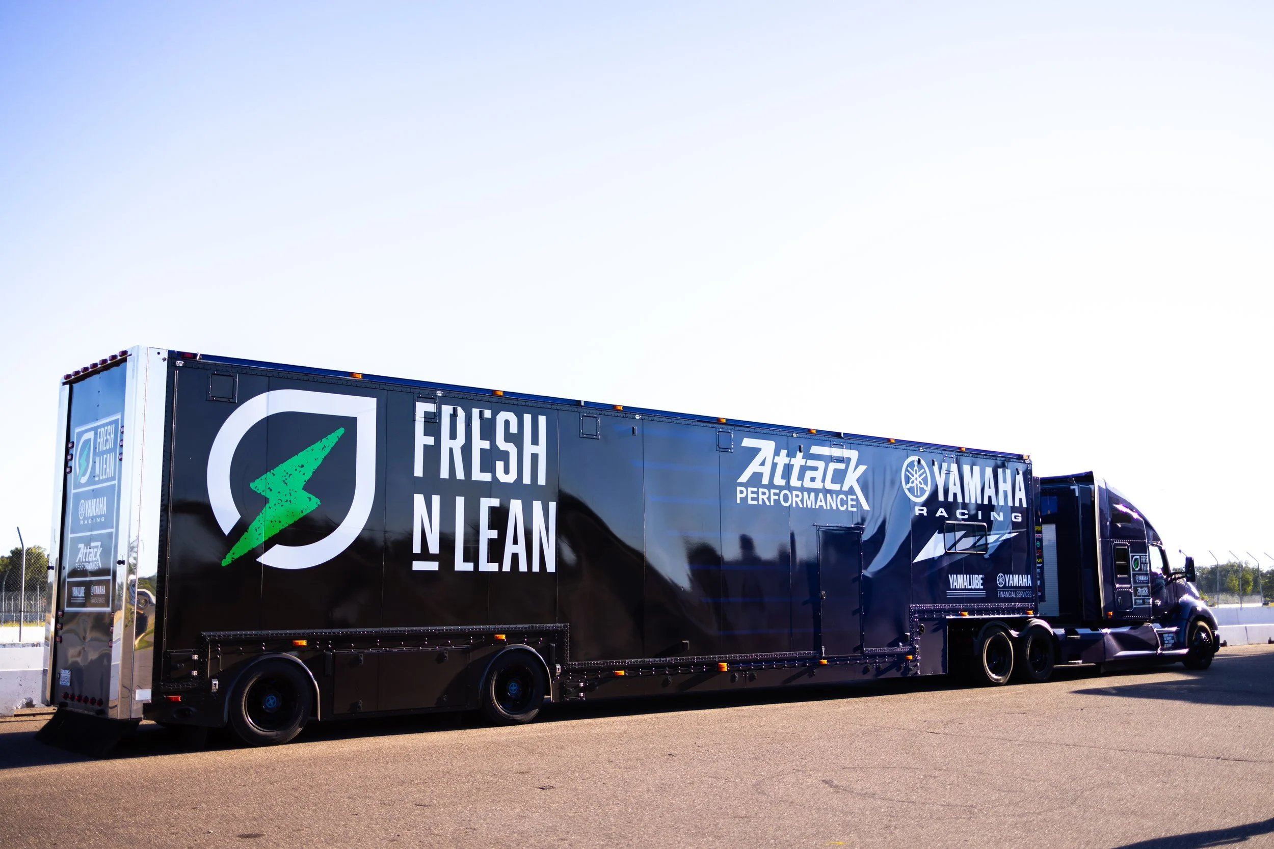 A large racing truck on a race track with promotional logos, including 'Fresh N Lean,' 'Attack Performance,' and 'Yamaha Racing,' parked on the asphalt under a clear sky.