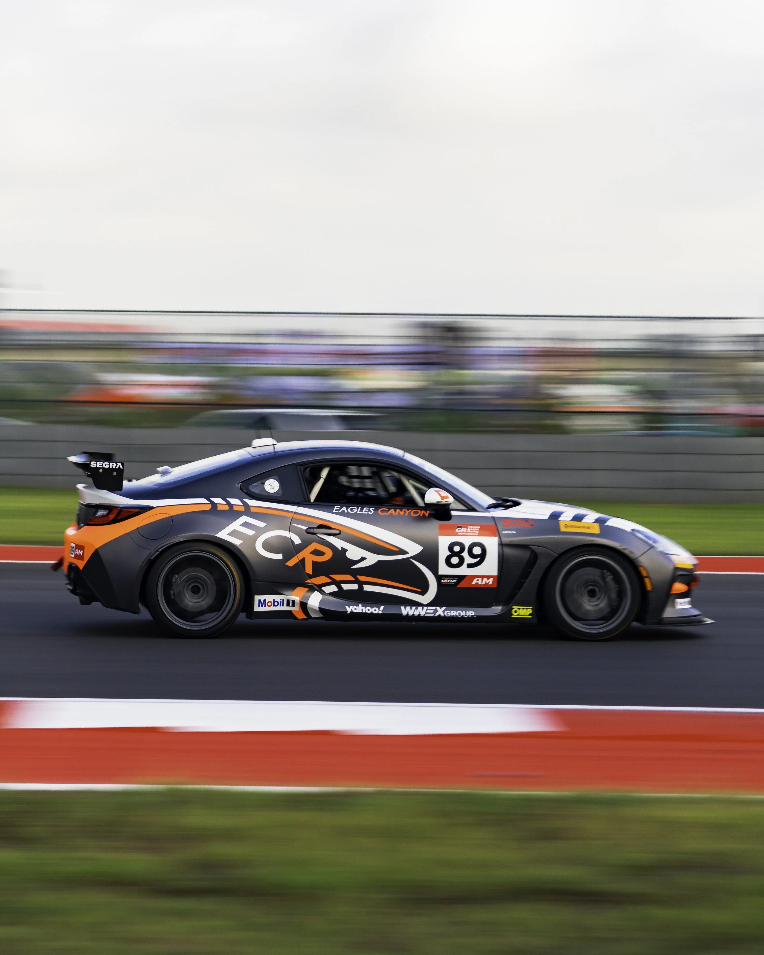 A race car with black, white, orange, and gray graphics, number 89, speeding on a race track with a blurred background.