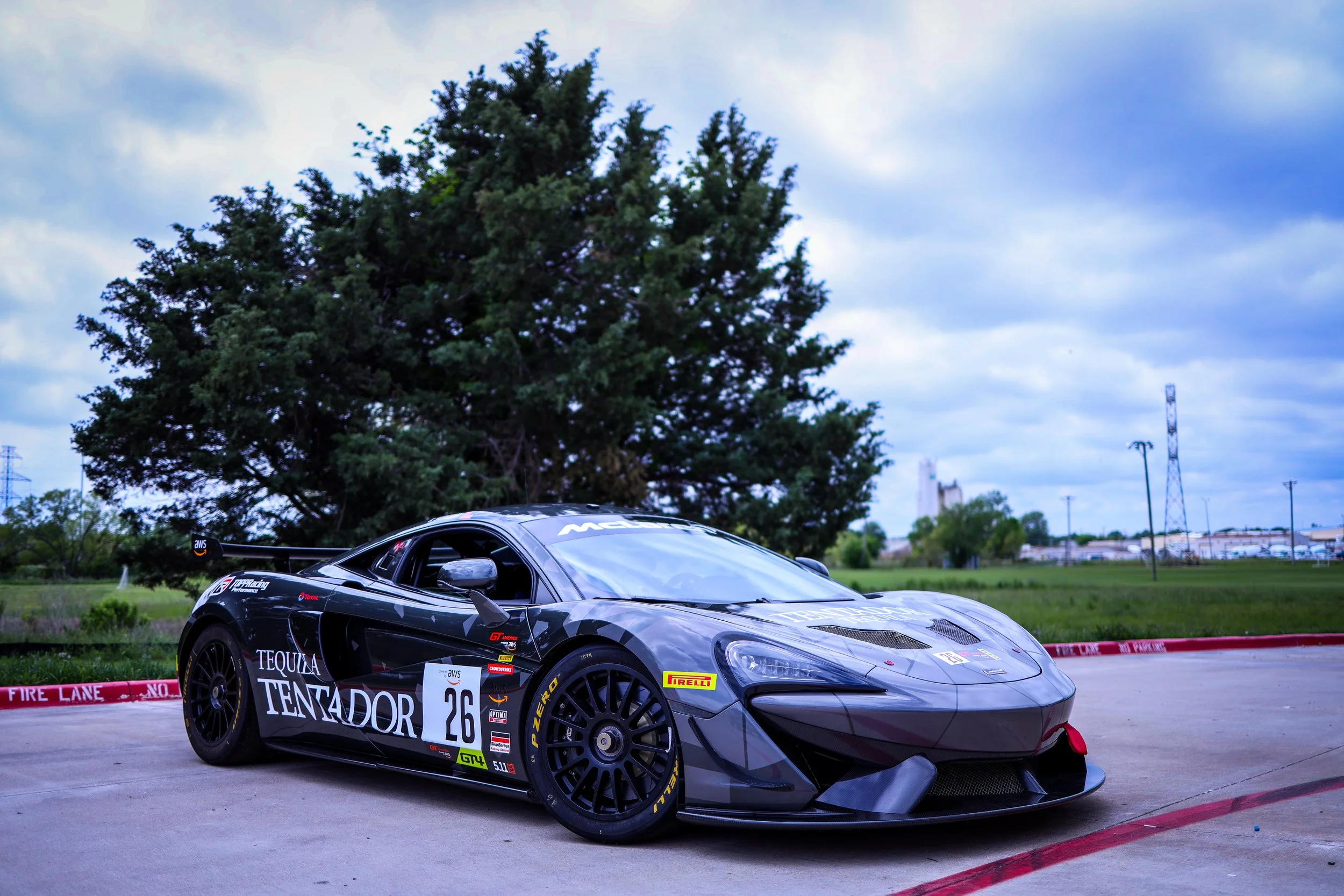 A black race car with the number 26 on its side, parked on a concrete surface next to a curb with a 'FIRE LANE NO PARKING' sign, with a large green tree and cloudy sky in the background.