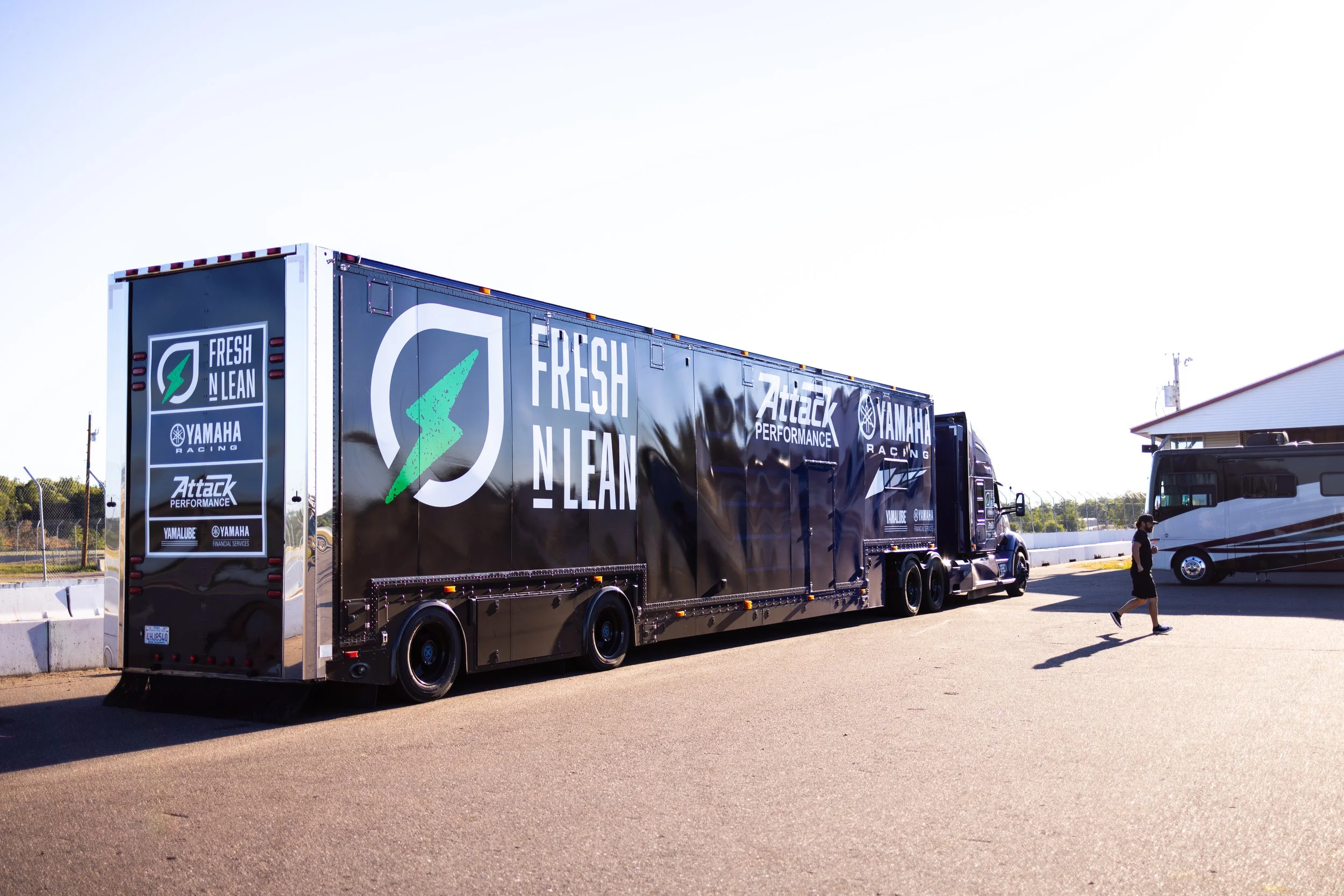Large black racing trailer with logos and branding for Fresh N Lean, Yamaha Racing, Attack Performance, and Yamalube, parked on a paved area near a racetrack. A man walks nearby, and there is a motorhome in the background.