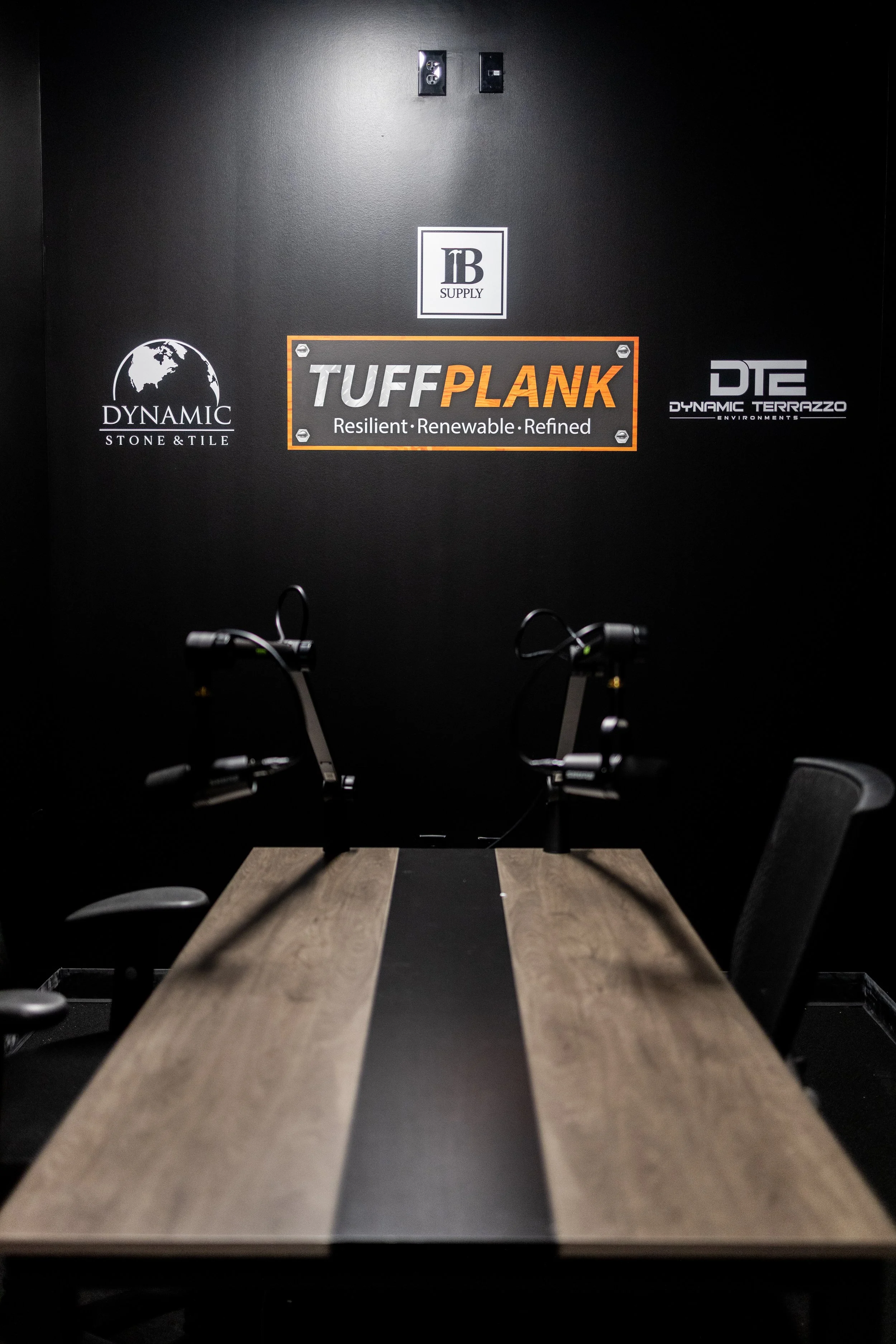 A conference room with a wooden table, two black office chairs, and two exercise machines mounted on the table. Graffiti style logos on the black wall in the background, including Turbo Plank, Dynamic Stone & Tile, Dynamic Terrazzo, and a logo with t
