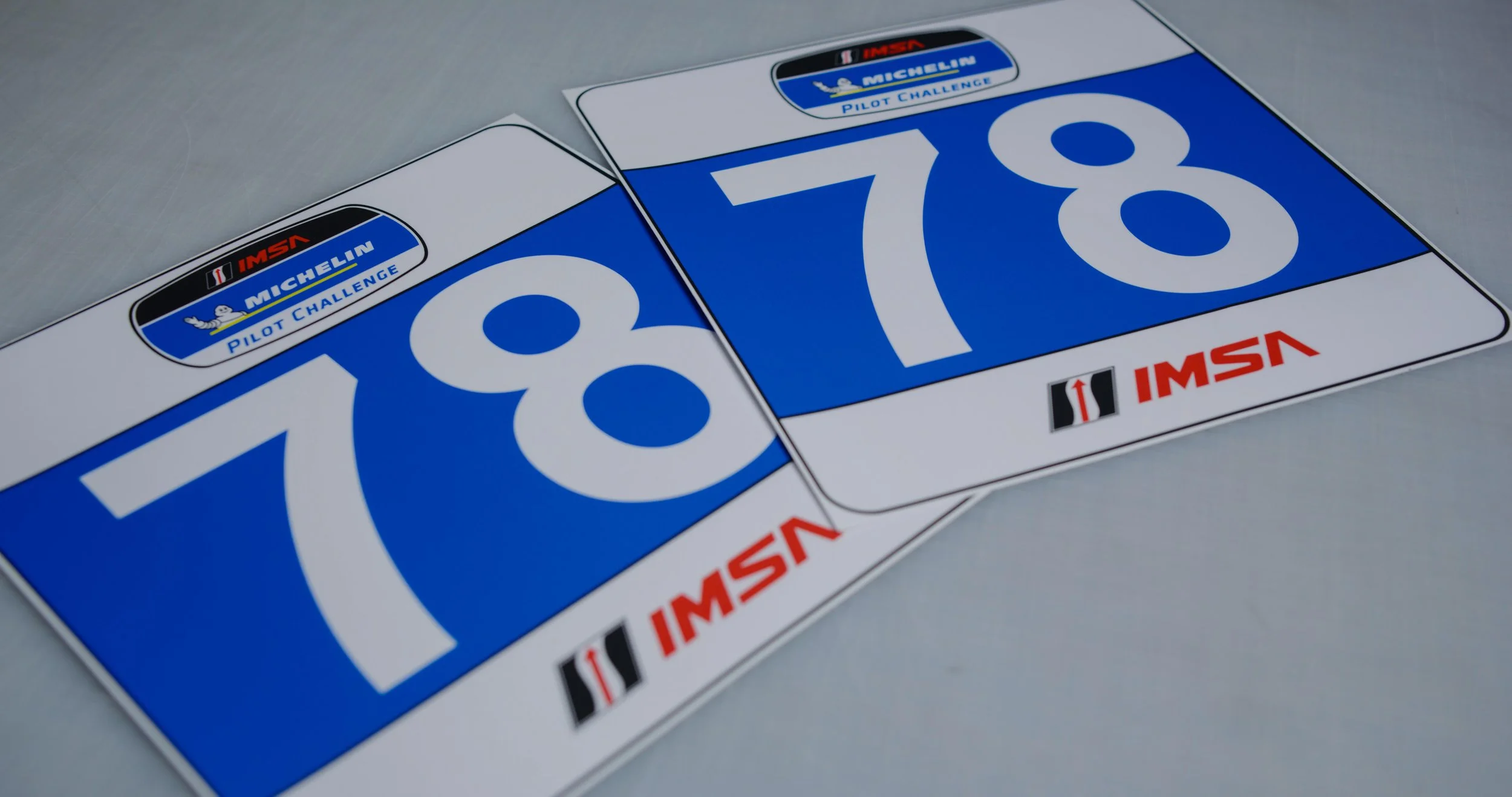 Two digital racing bibs with blue backgrounds and white numbers, 78 and 78, laid flat on a gray surface, featuring IMSA and Michelin logos.