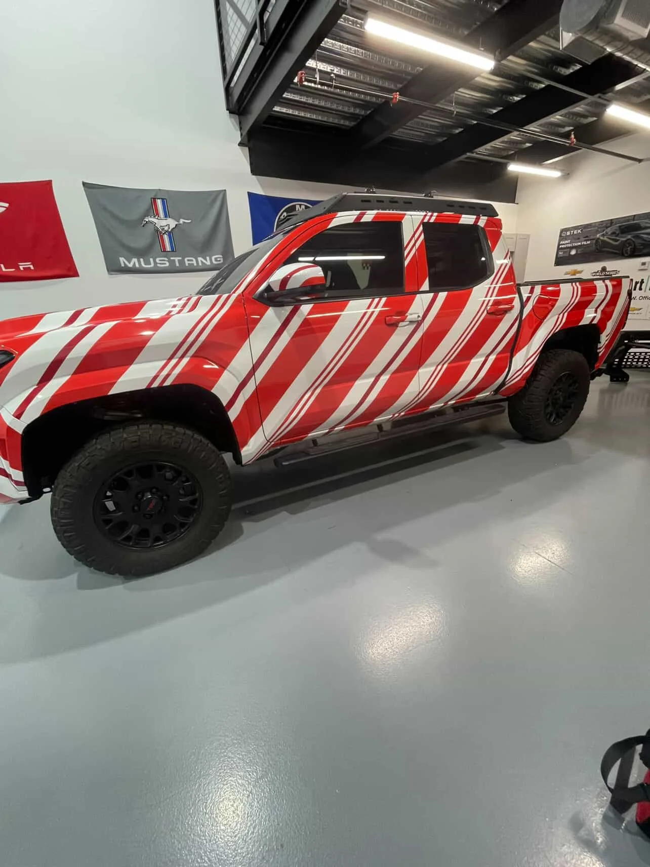 A red pickup truck with white diagonal stripe wrap parked indoors, with Mustang and other car brand flags on the wall behind it.