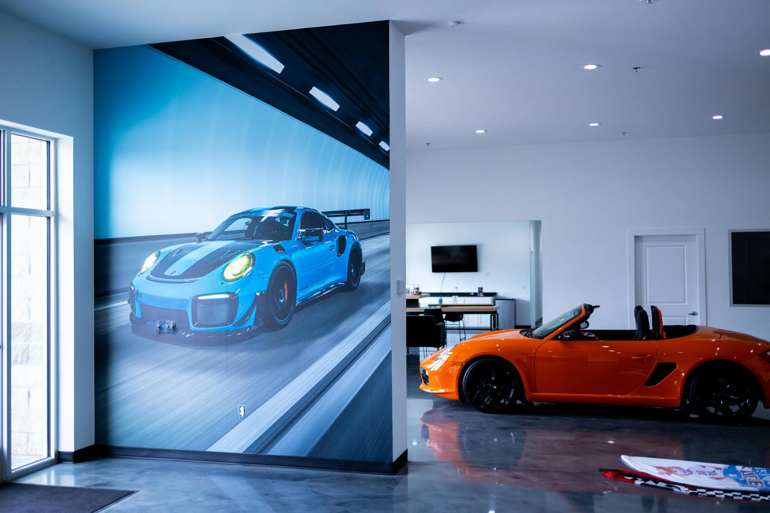 Interior of a car showroom with two sports cars: an orange convertible and a blue racing car on a large wall mural.