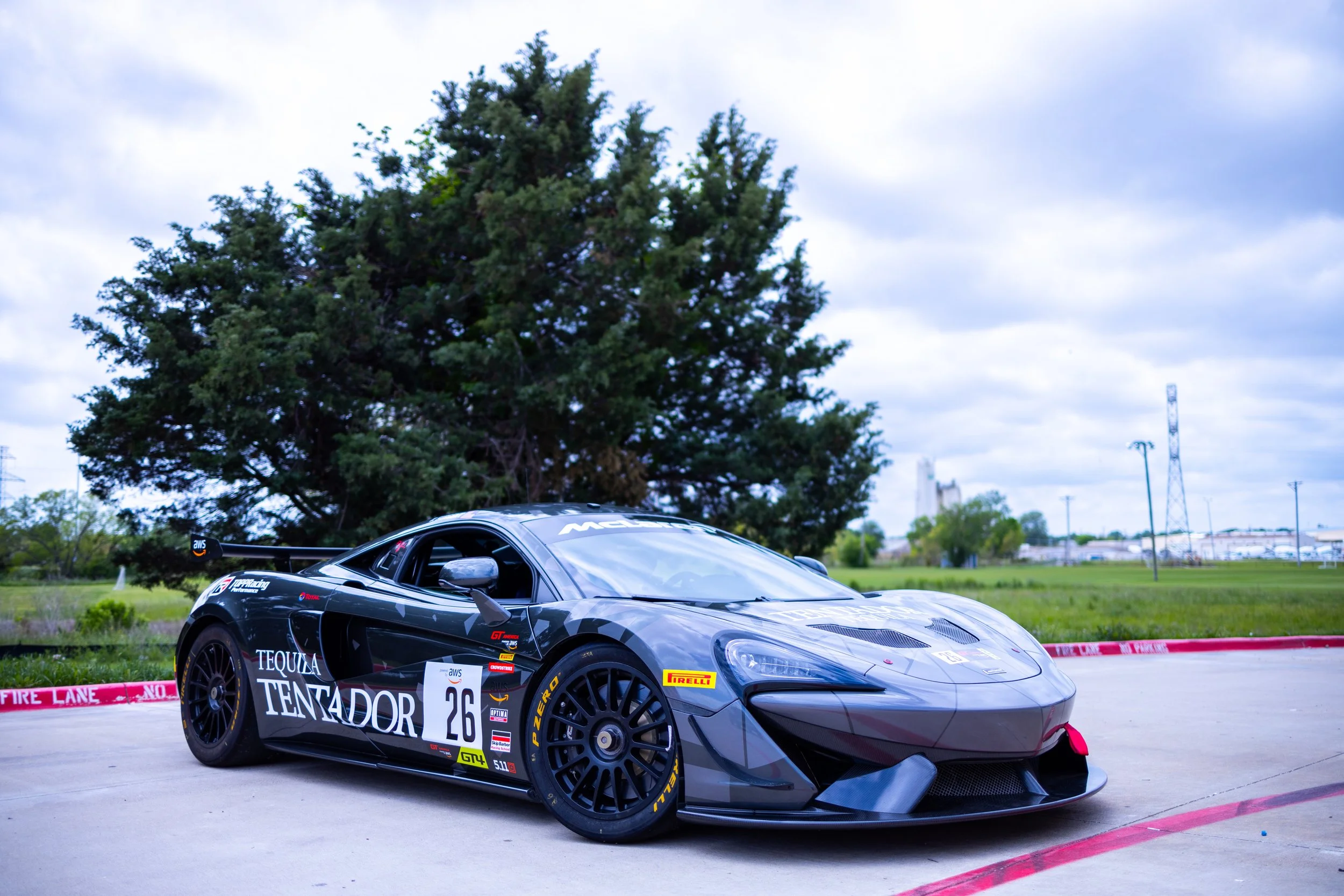 A racing car with the number 26 parked on a concrete surface near a green grassy area and trees under a partly cloudy sky.