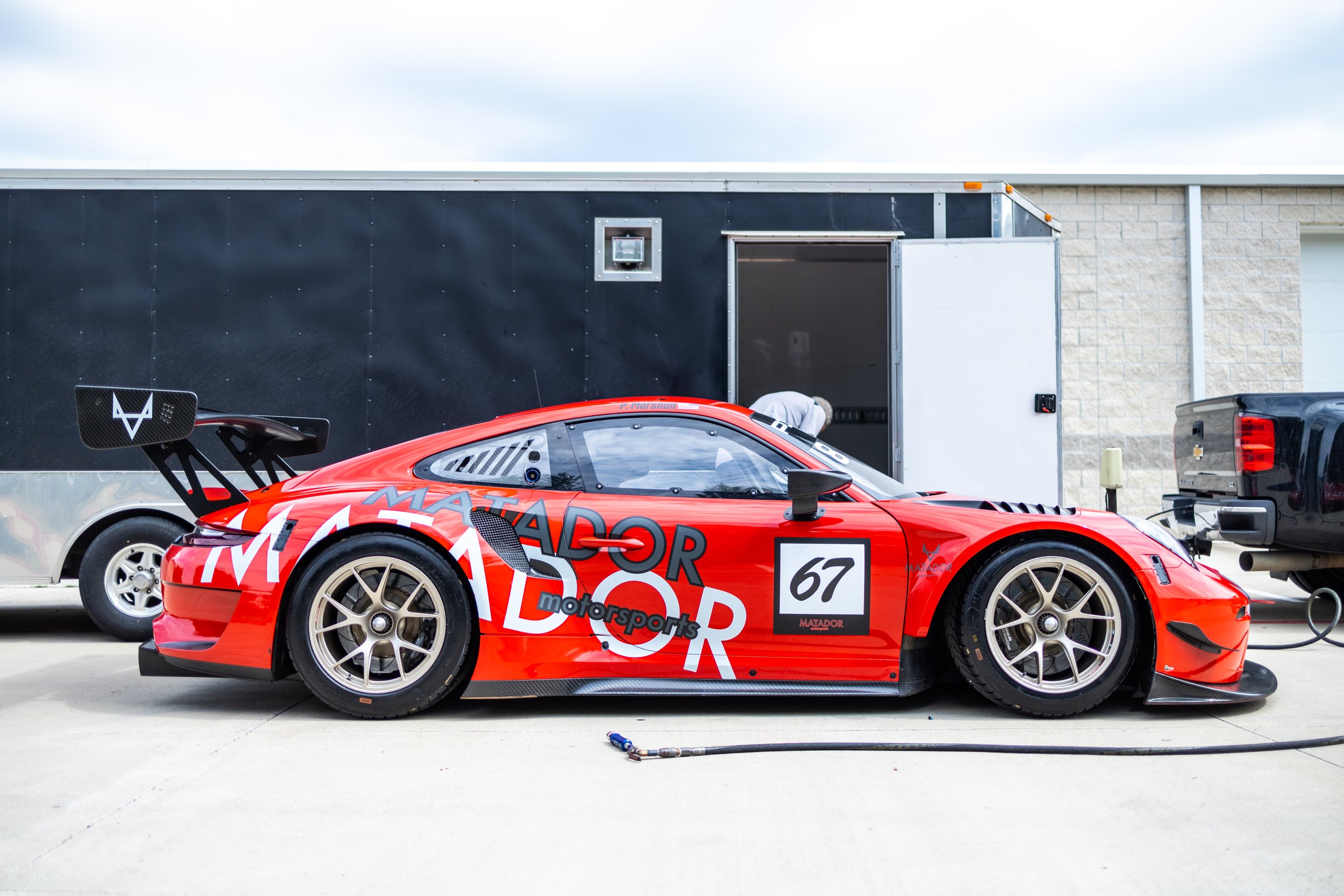 Red race car with black rear wing and white decals, parked outside a building, connected to a black pickup truck for refueling or maintenance.