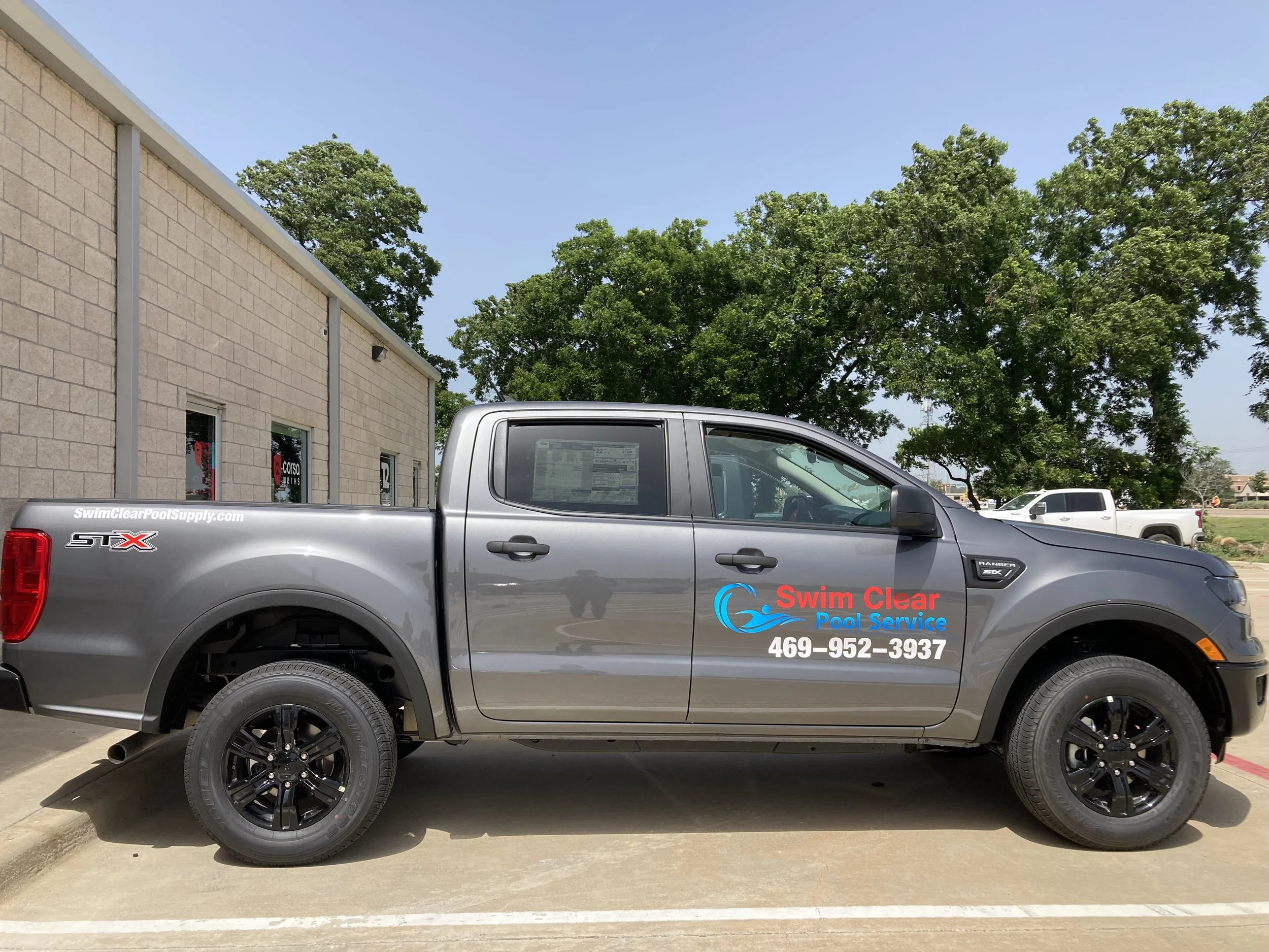 Gray pickup truck parked outside a building, with a sign on the side that reads 'Swim Clear Pool Service' along with a phone number, 469-952-3937. The truck has black wheels and branding for pool services.