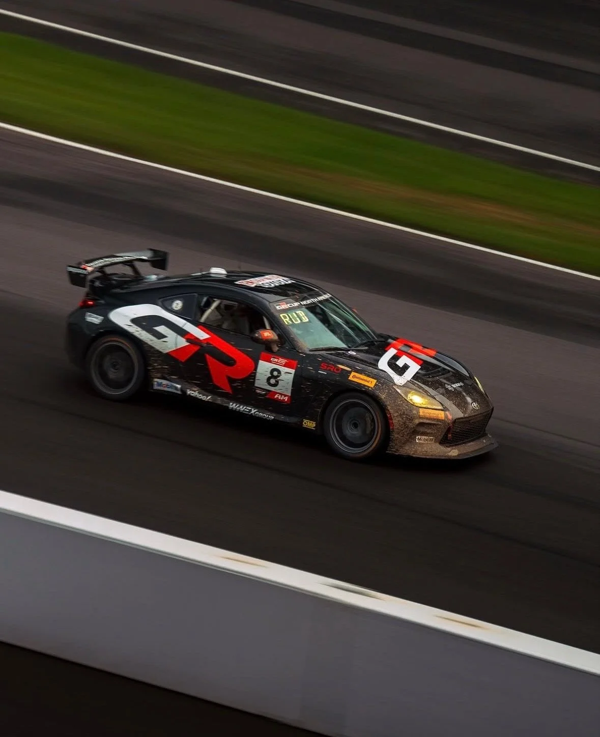 A black race car with red and white racing decals driving on a racetrack, background showing blurred asphalt and green grass.