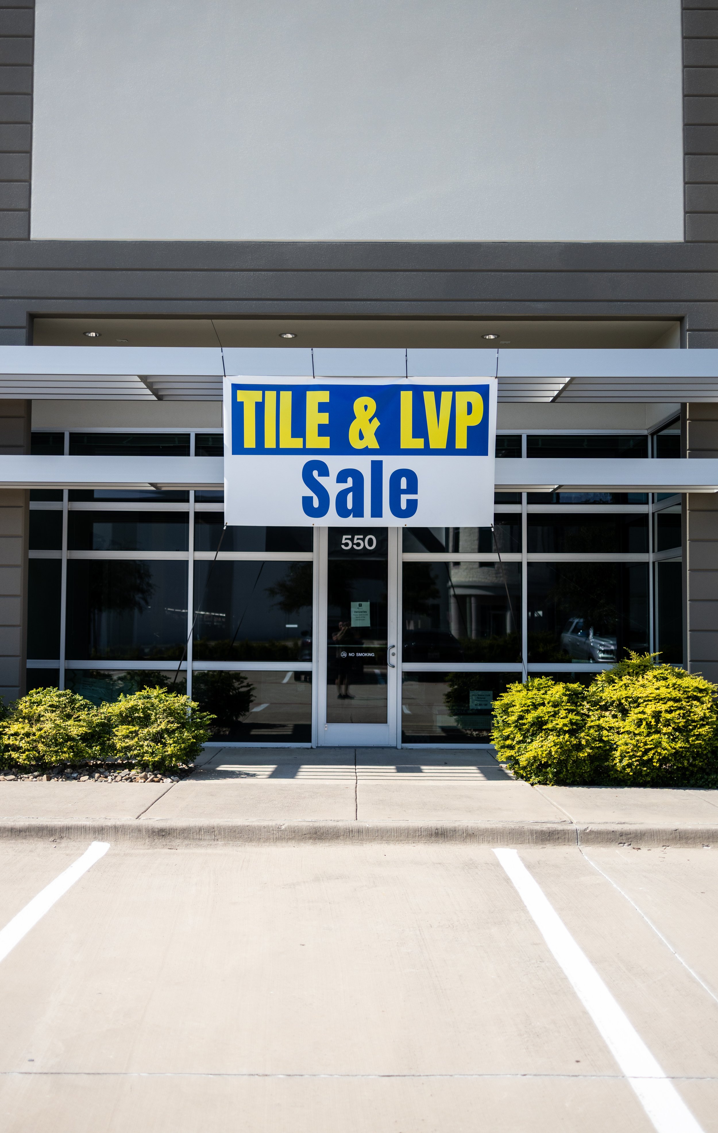 A storefront with a large sign announcing a tile and LVP sale, located at 550, with small bushes on either side of the entrance and parking spaces in front.