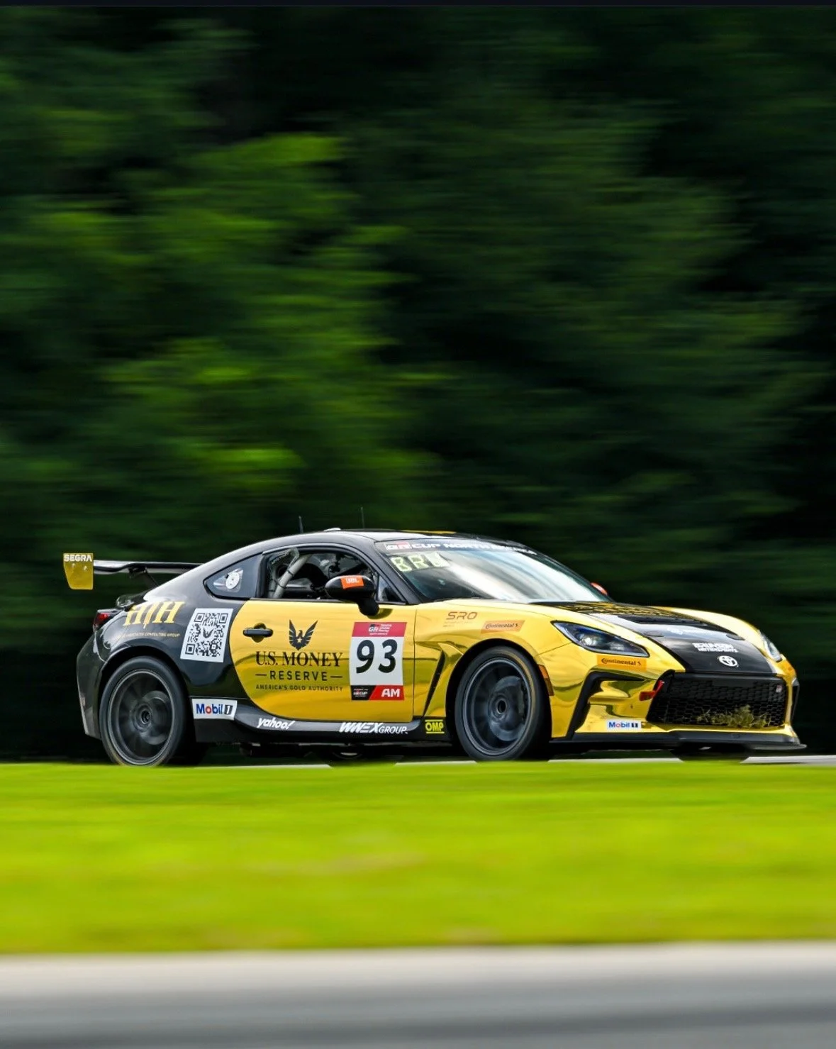 A yellow and black race car with the number 93 is speeding on a track, with a blurred green foliage background.