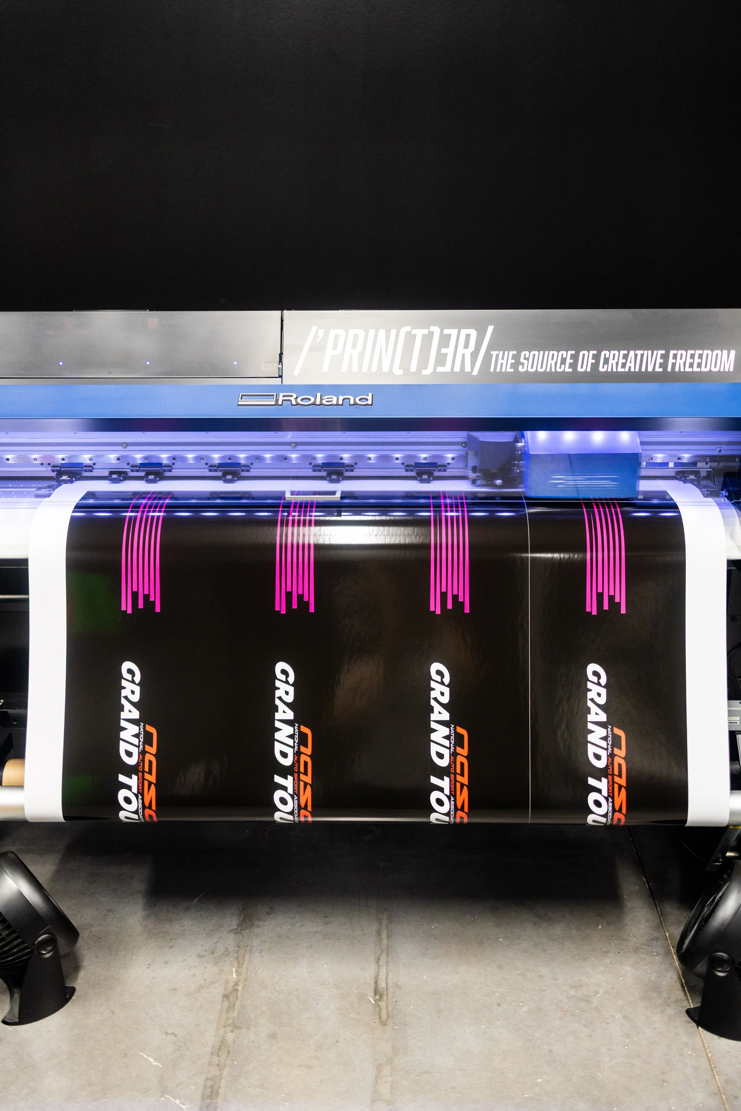 Large format printer printing a banner with the text 'GRAND TOUR' and 'MSD' in bold letters, with black, white, and orange colors.