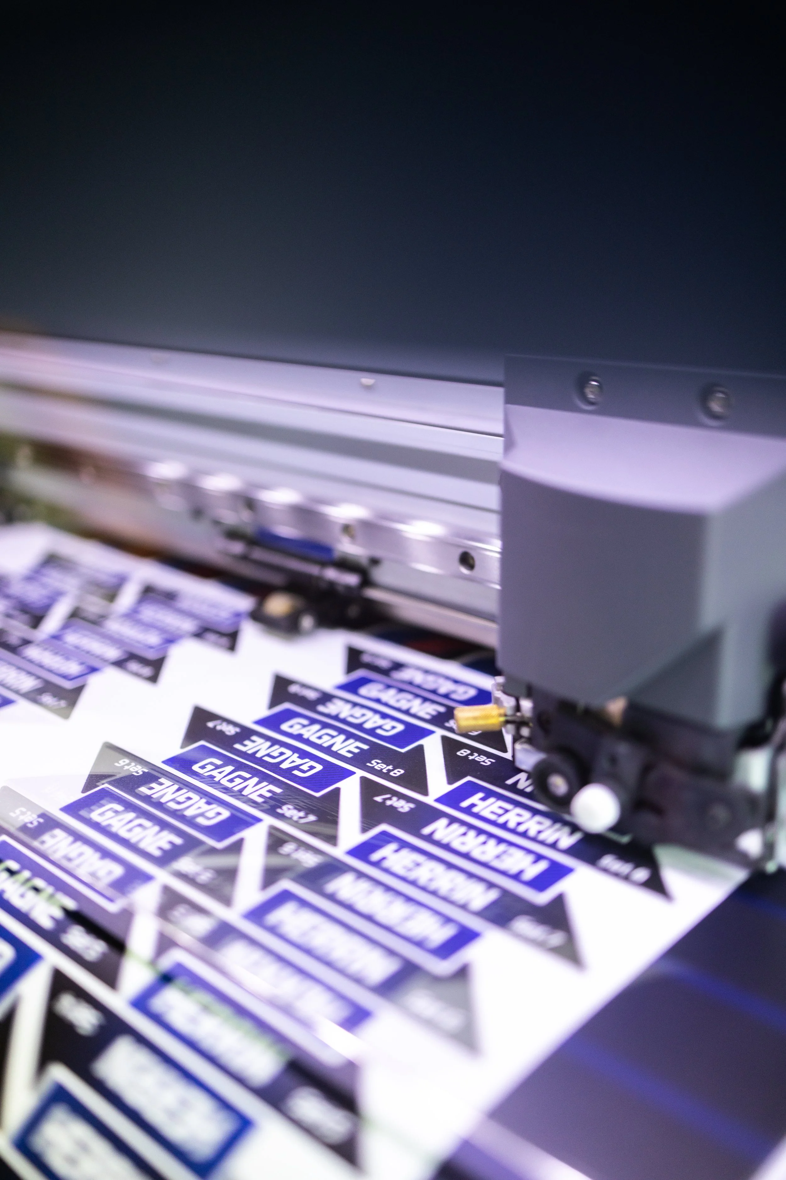 A printing machine is producing sheets of stickers with the name "GAGNE" in white text on a blue background and "HERRIN" in white text on a black background.