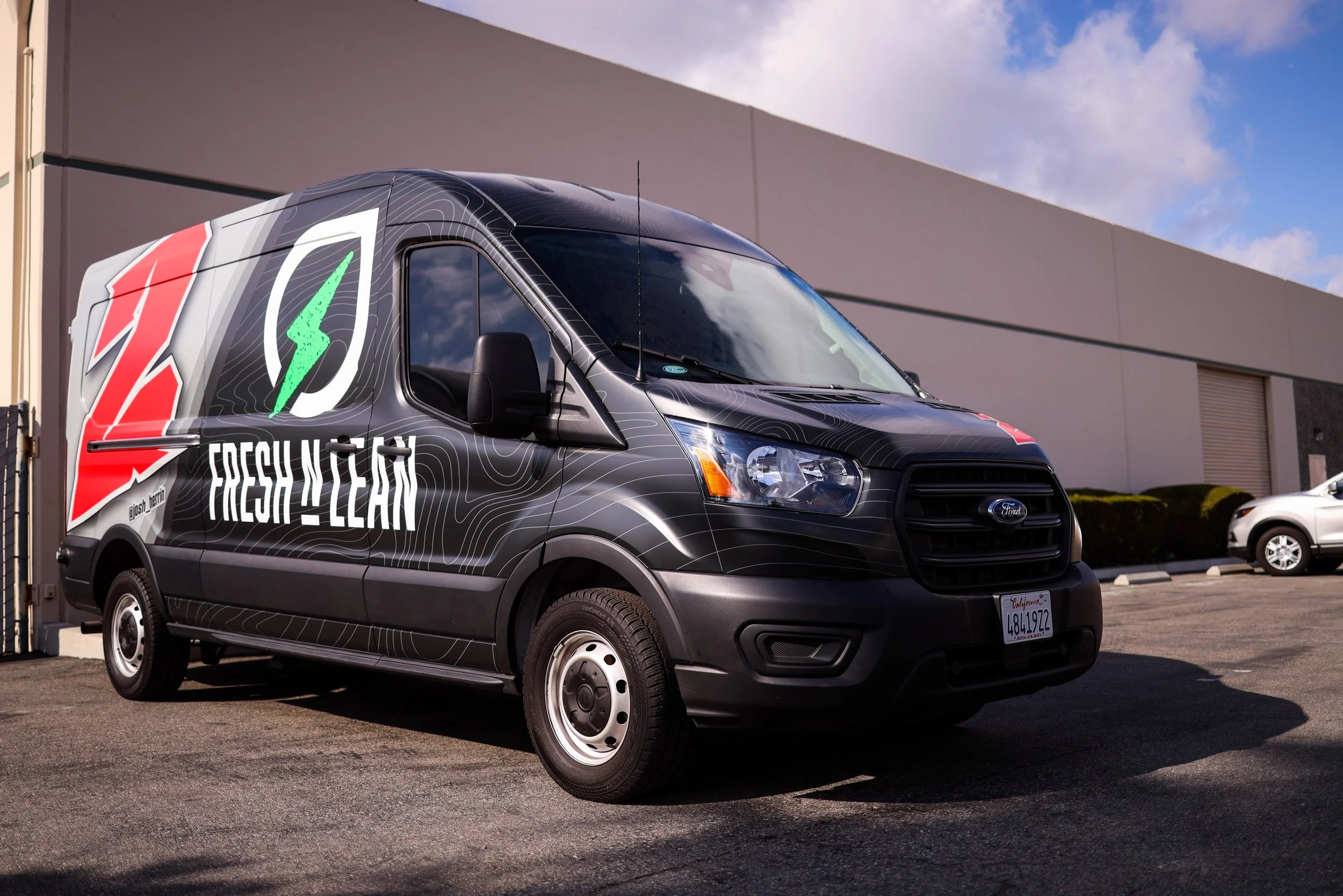 Black delivery van with promotional graphics that reads 'Fresh n Lean,' parked in a lot next to a modern industrial building.