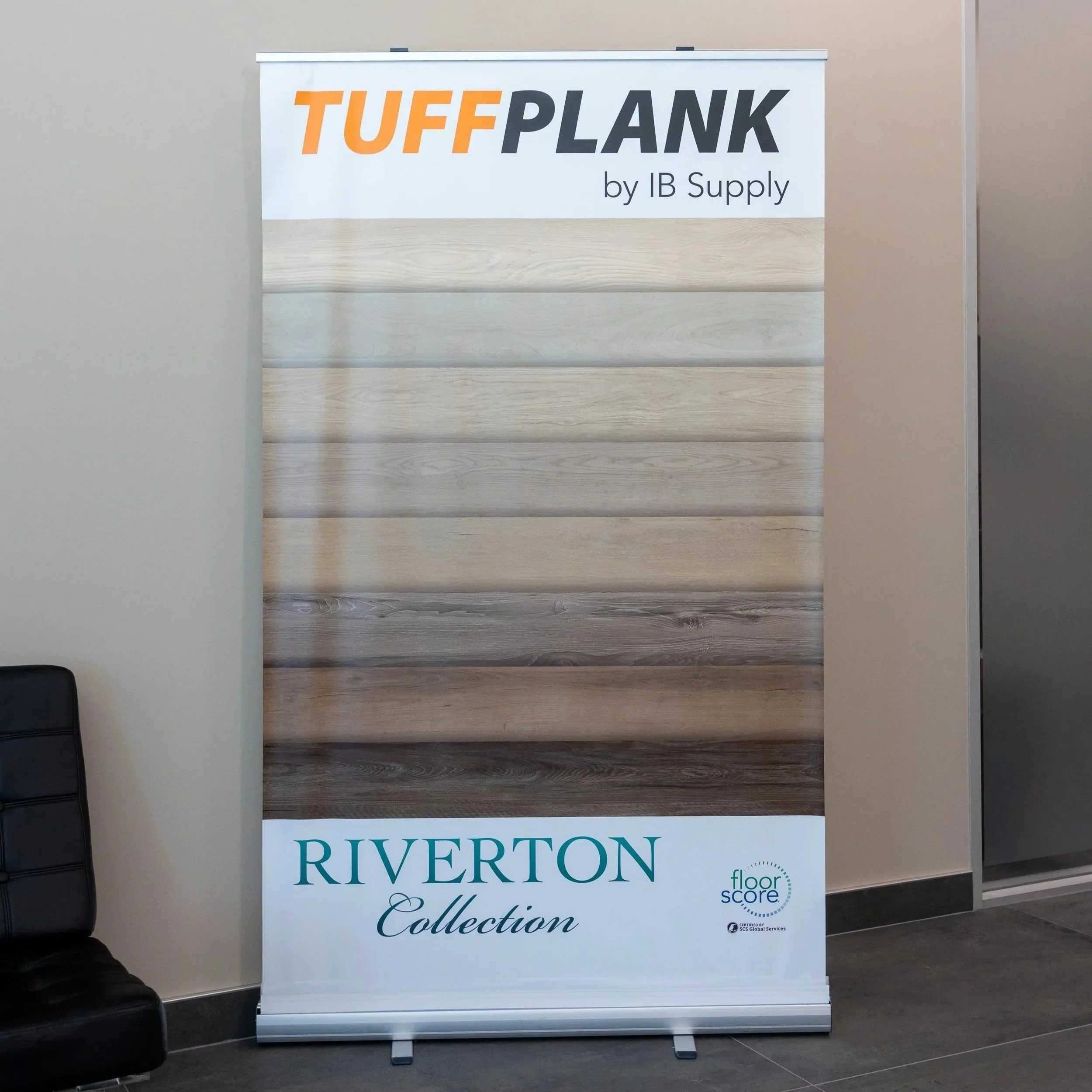 Display stand showcasing various wood flooring samples from the Riverton Collection by TUFFPLANK, IB Supply, with a white top and bottom section, placed indoors near a wall and a black chair.