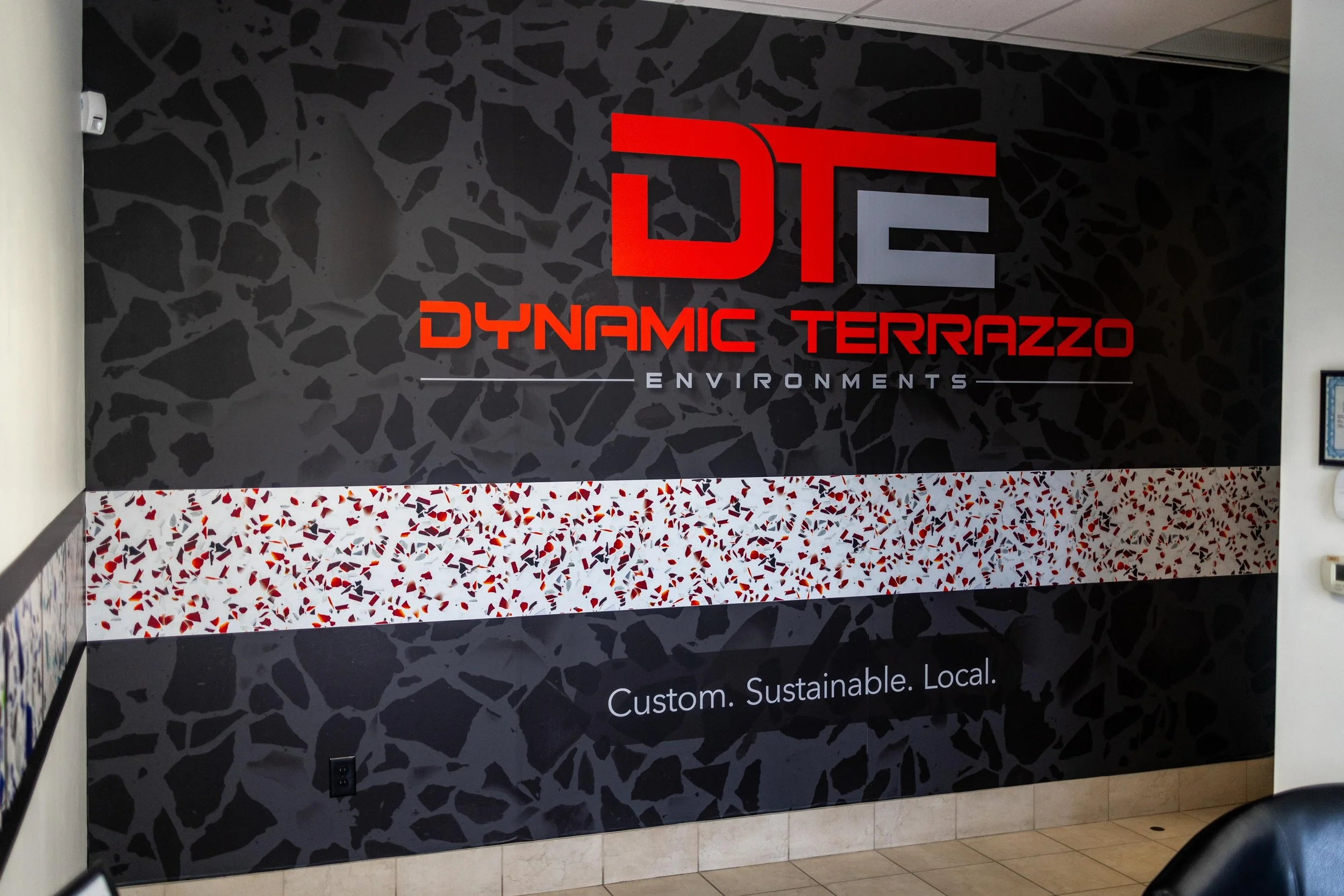 Wall with logo and text for Dynamic Terrazzo Environments. The logo features large red and gray letters DTE, with the words "Dynamic Terrazzo" in red below the logo, and "Environments" in smaller gray text beneath. The wall has a black, terrazzo-style pattern with a horizontal strip of colorful terrazzo pattern in the middle. The tagline at the bottom reads "Custom. Sustainable. Local."