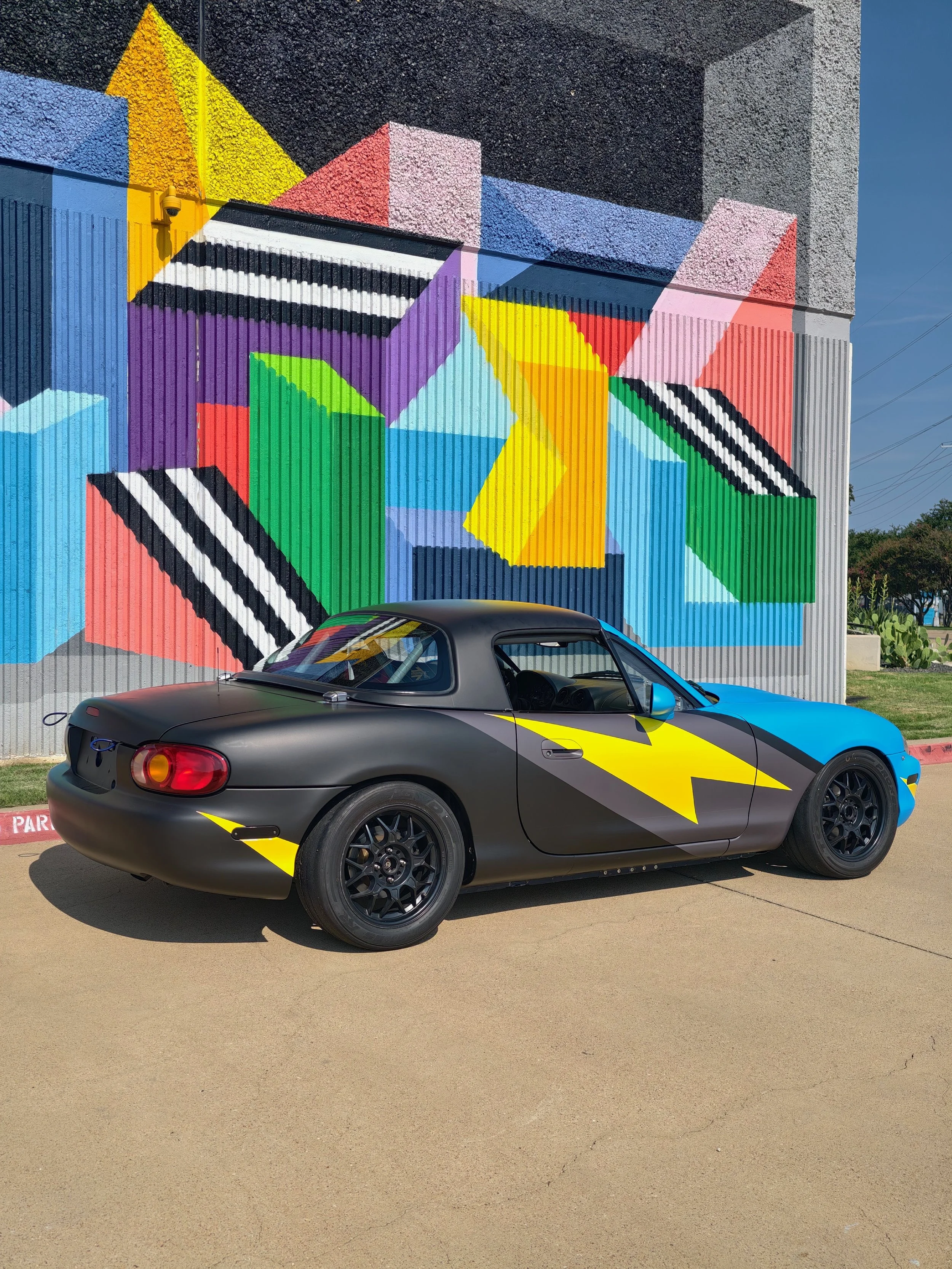 A sports car painted with black, blue, and yellow lightning bolt designs parked in front of a colorful geometric mural on a wall