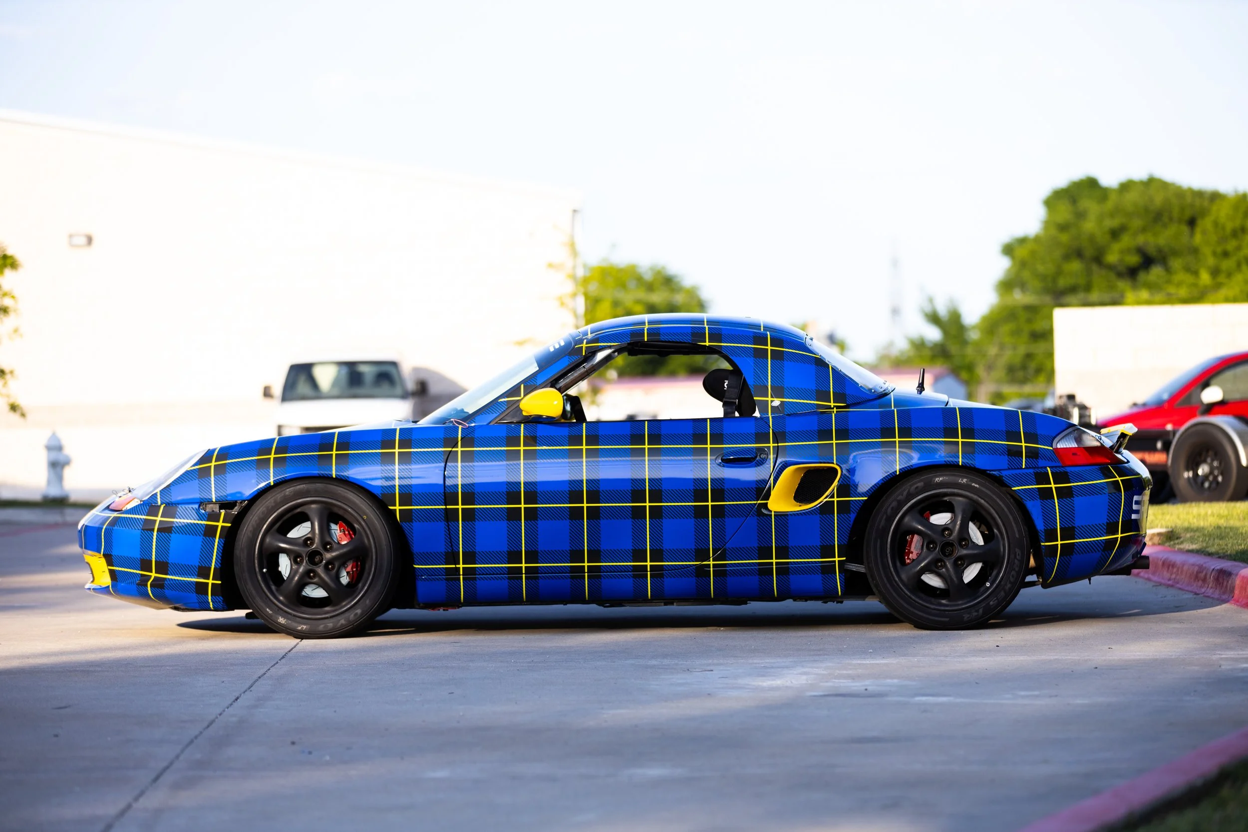 A blue sports car with a yellow checkered pattern parked on a concrete surface, with a white building and trees in the background.