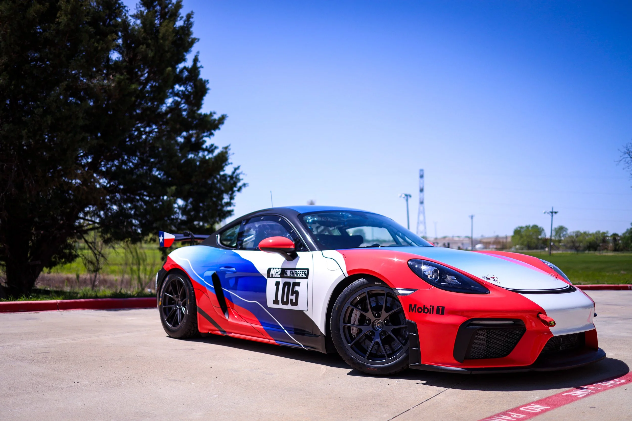 A race car painted in red, white, and blue, parked on a paved area with trees and a clear blue sky in the background.
