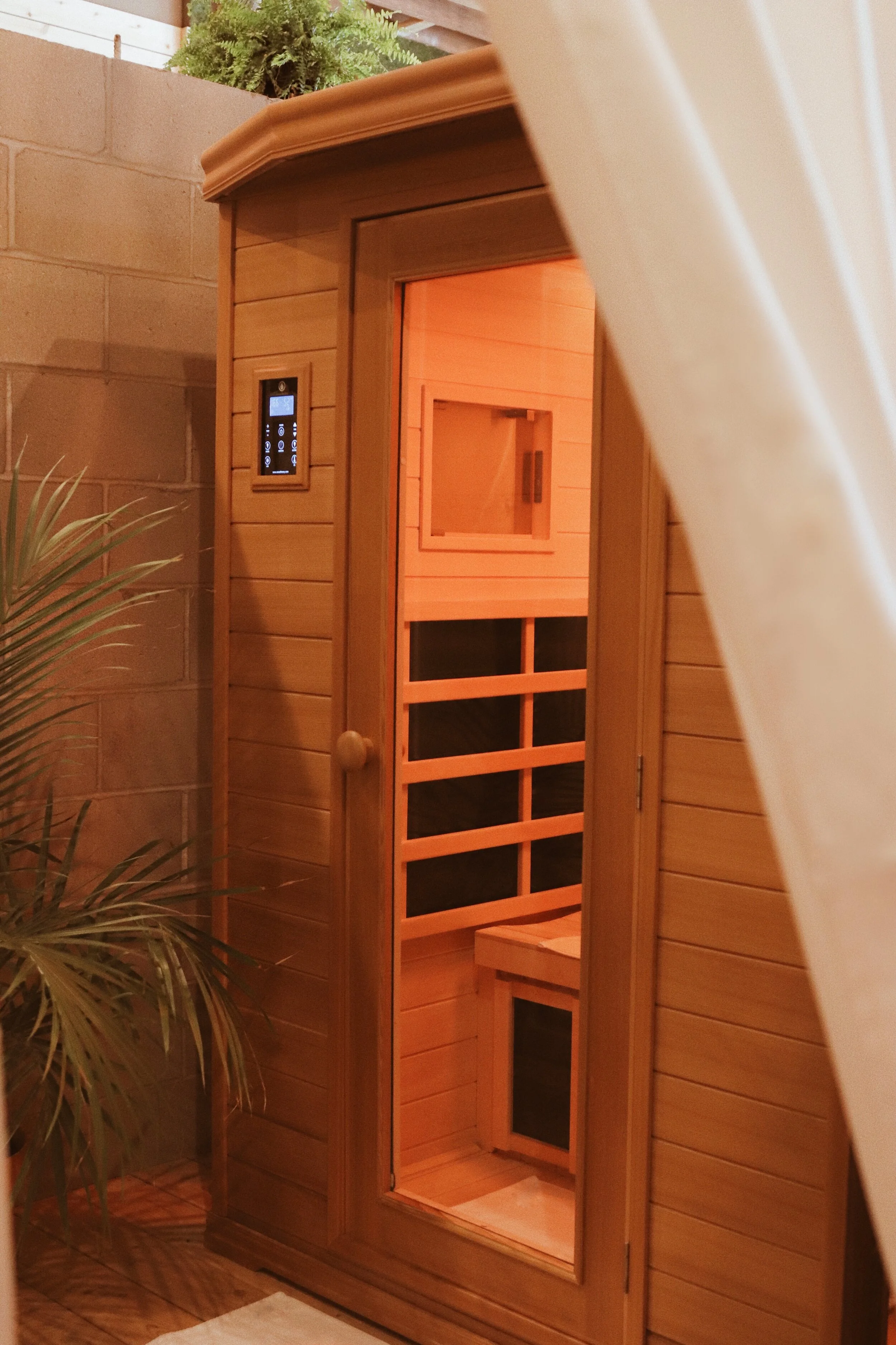 A wooden infrared sauna with a control panel on the exterior. The interior has wooden benches and a back wall with glass panels, illuminated by orange-red light. An indoor plant is partially visible o