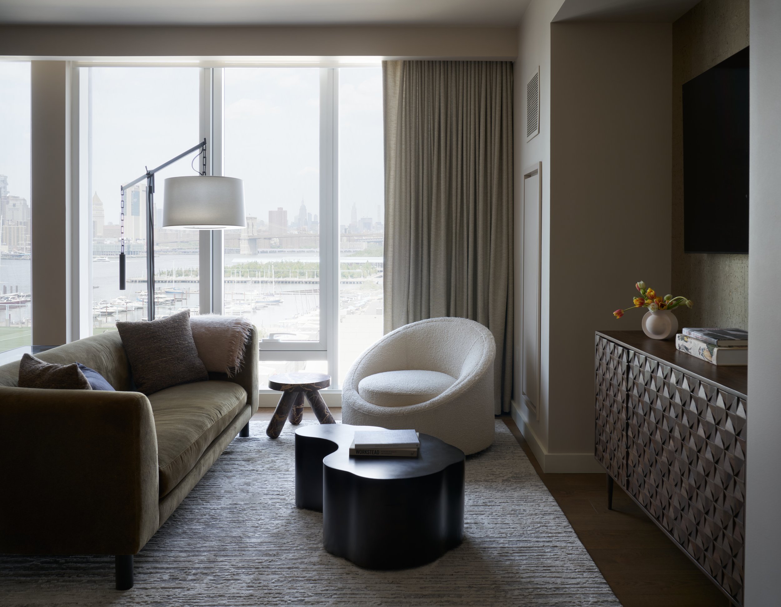 Modern living room with large windows overlooking a cityscape and river, beige sofa with pillows, white rounded chair, black coffee table, small wooden side table, tall floor lamp, patterned sideboard with a vase of flowers, books, and a wall-mounted