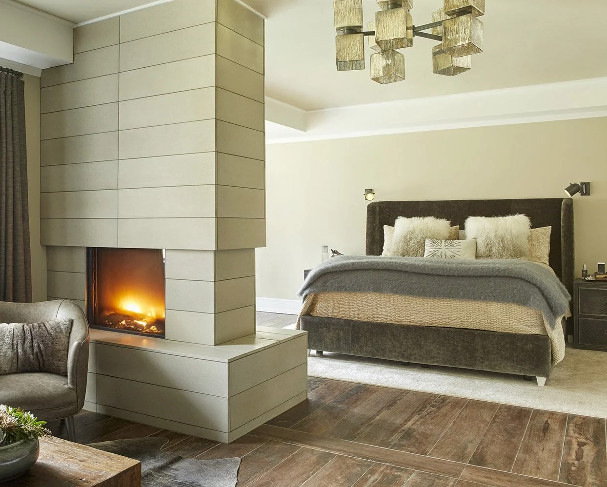 Modern bedroom with a bed, plush pillows, and a fireplace, featuring beige walls, dark wood flooring, and contemporary lighting fixtures.