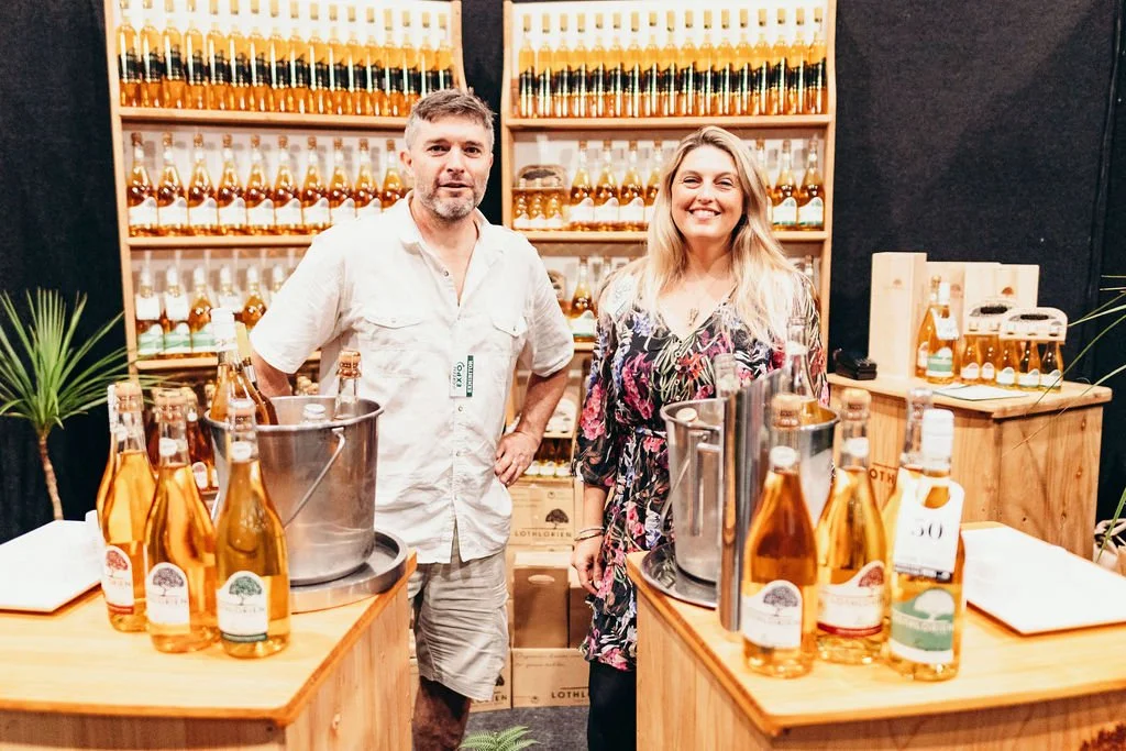 A man and a woman stand behind a wooden table displaying bottles of amber-colored liquids, likely honey or syrup, at a market stall with a wooden shelving unit filled with similar bottles in the background.