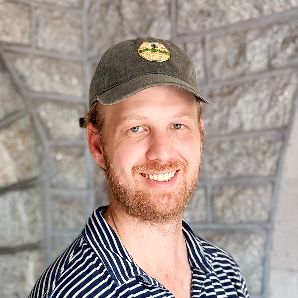 A young man with a beard, wearing a striped shirt and a cap, smiling in front of a stone wall background.