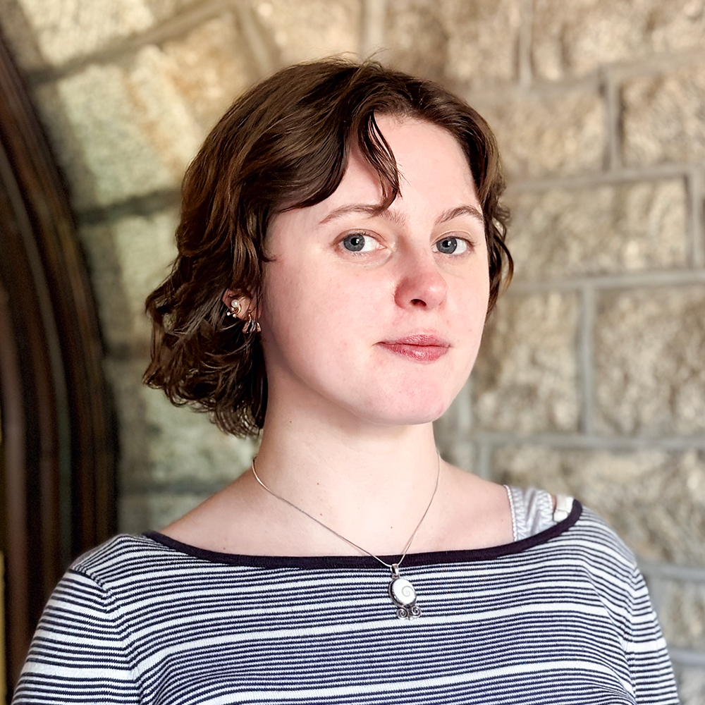 A young woman with short wavy brown hair, blue eyes, and fair skin standing outdoors in front of a stone wall, wearing a striped shirt and a silver necklace.