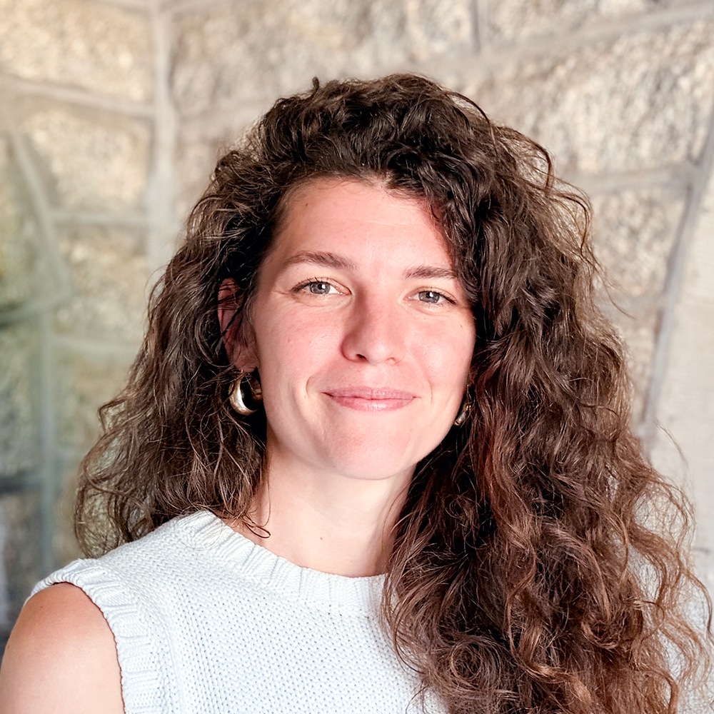 A woman with curly brown hair, earrings, and a white sleeveless top stands in front of a stone wall.