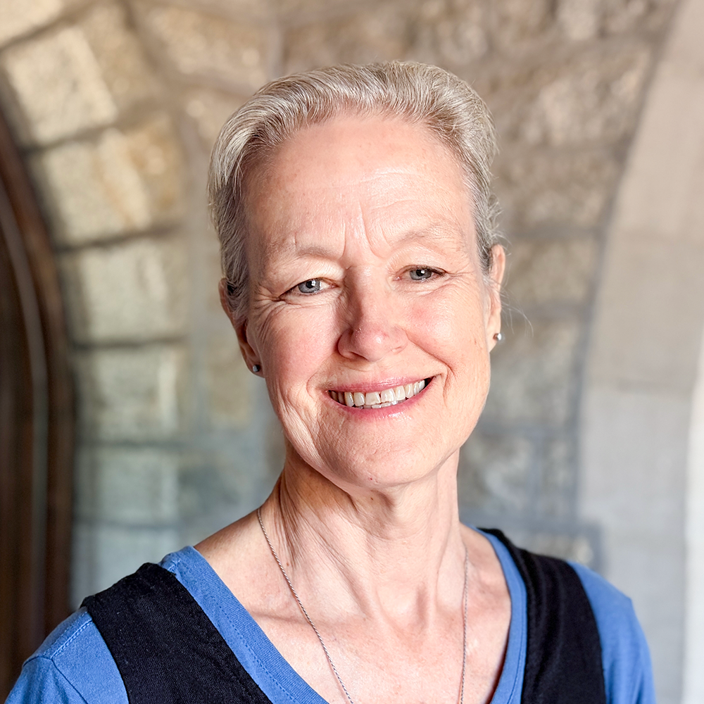 Close-up of a smiling older woman with short silver hair, wearing a blue shirt and a black vest, standing outdoors against a stone wall background.