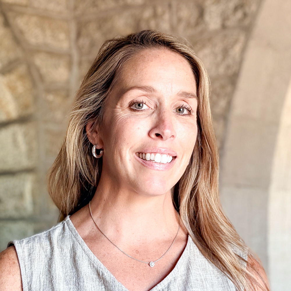 A woman with long blonde hair, wearing a light-colored sleeveless top, silver earrings, and a delicate necklace with a small pendant, stands in front of a stone archway and smiles at the camera.