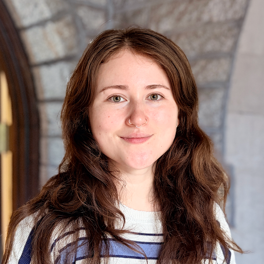A young woman with wavy brown hair, green eyes, and a septum piercing, smiling indoors with a stone fireplace in the background.