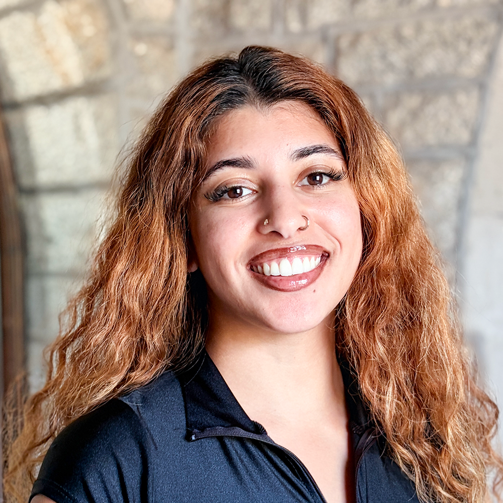 A woman with long, curly red hair, smiling in front of a brick wall, wearing a black zip-up top.