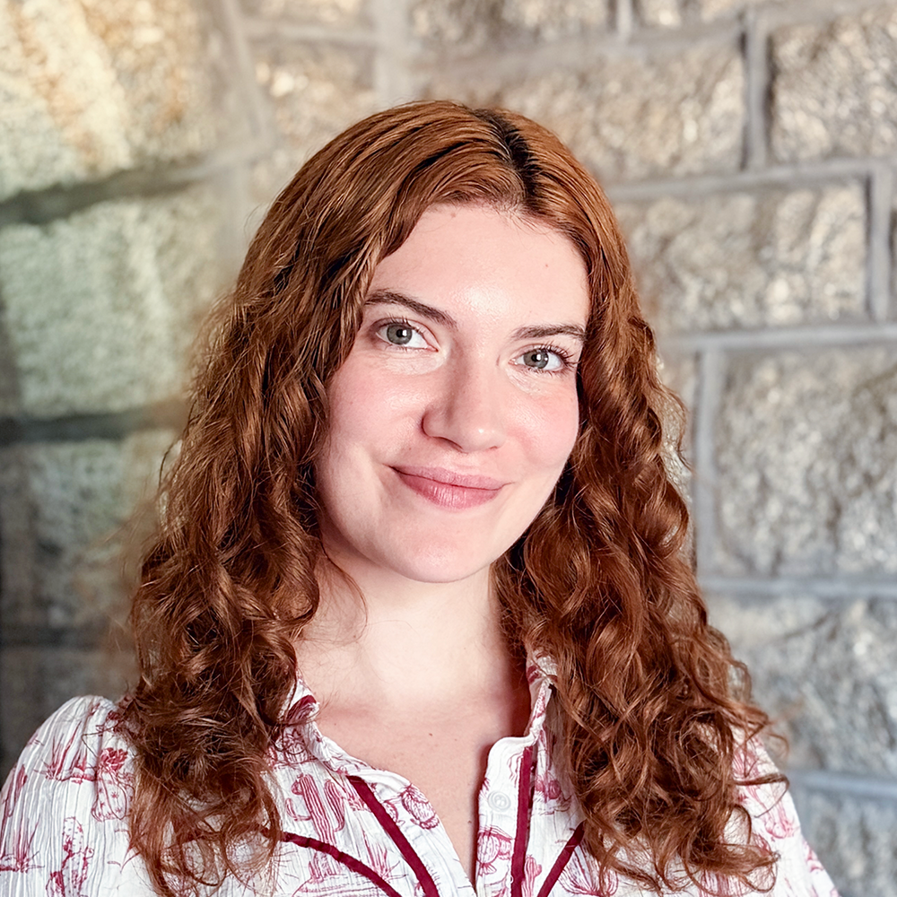 A woman with long, curly red hair smiling in front of a stone wall.