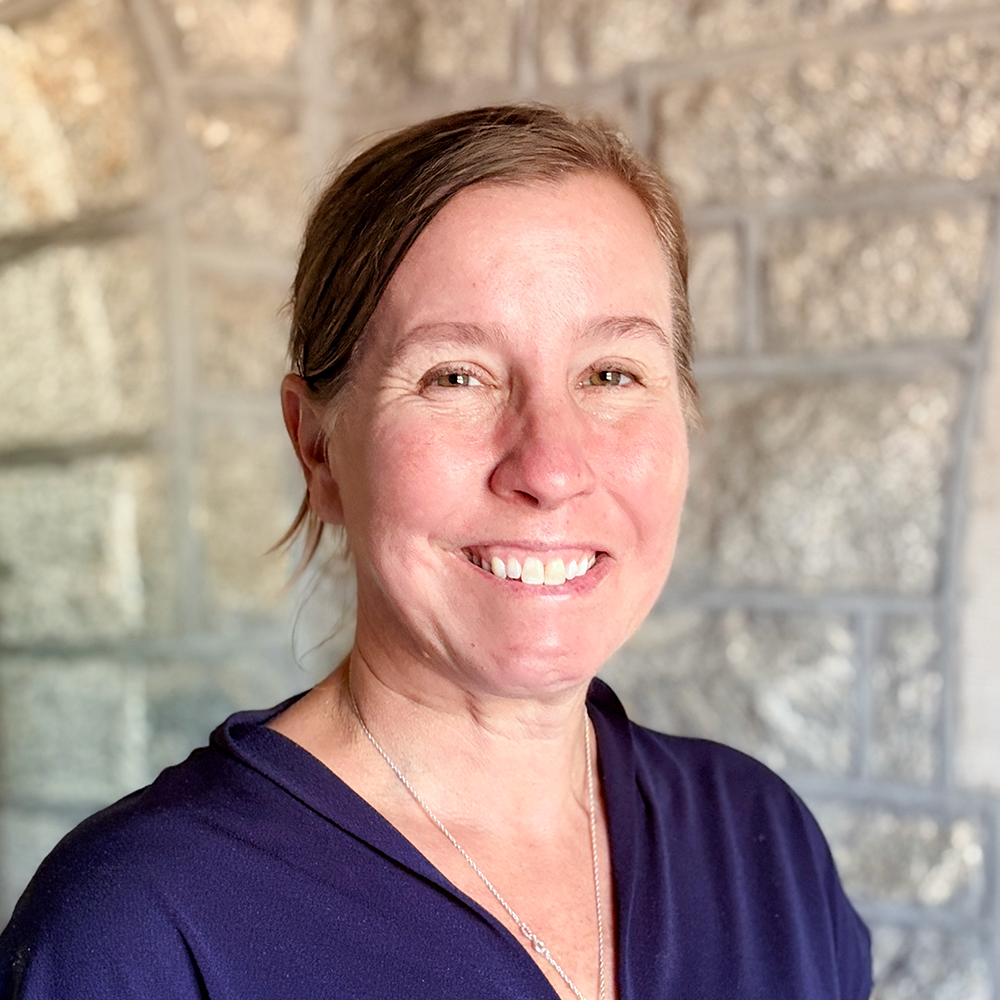 A woman smiling with short brown hair, wearing a navy blue top and a silver necklace, standing in front of a stone wall.