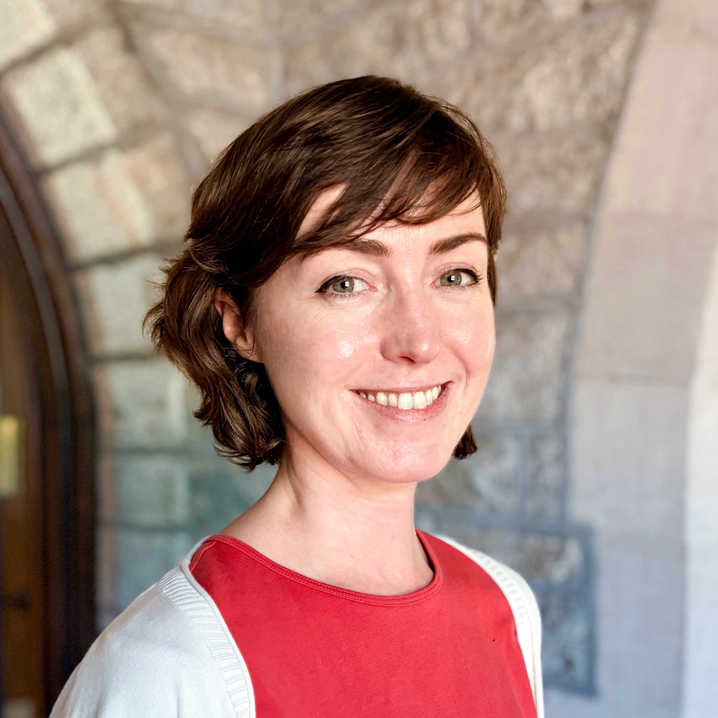 Close-up of a woman with short brown hair, green eyes, smiling, wearing a red and white top, standing against a blurred brick wall background.