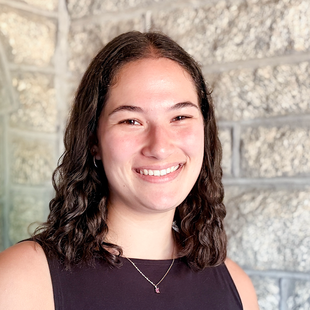 Portrait of a young woman with curly brown hair, smiling, wearing a black top and a sticking necklace, standing against a textured stone wall.