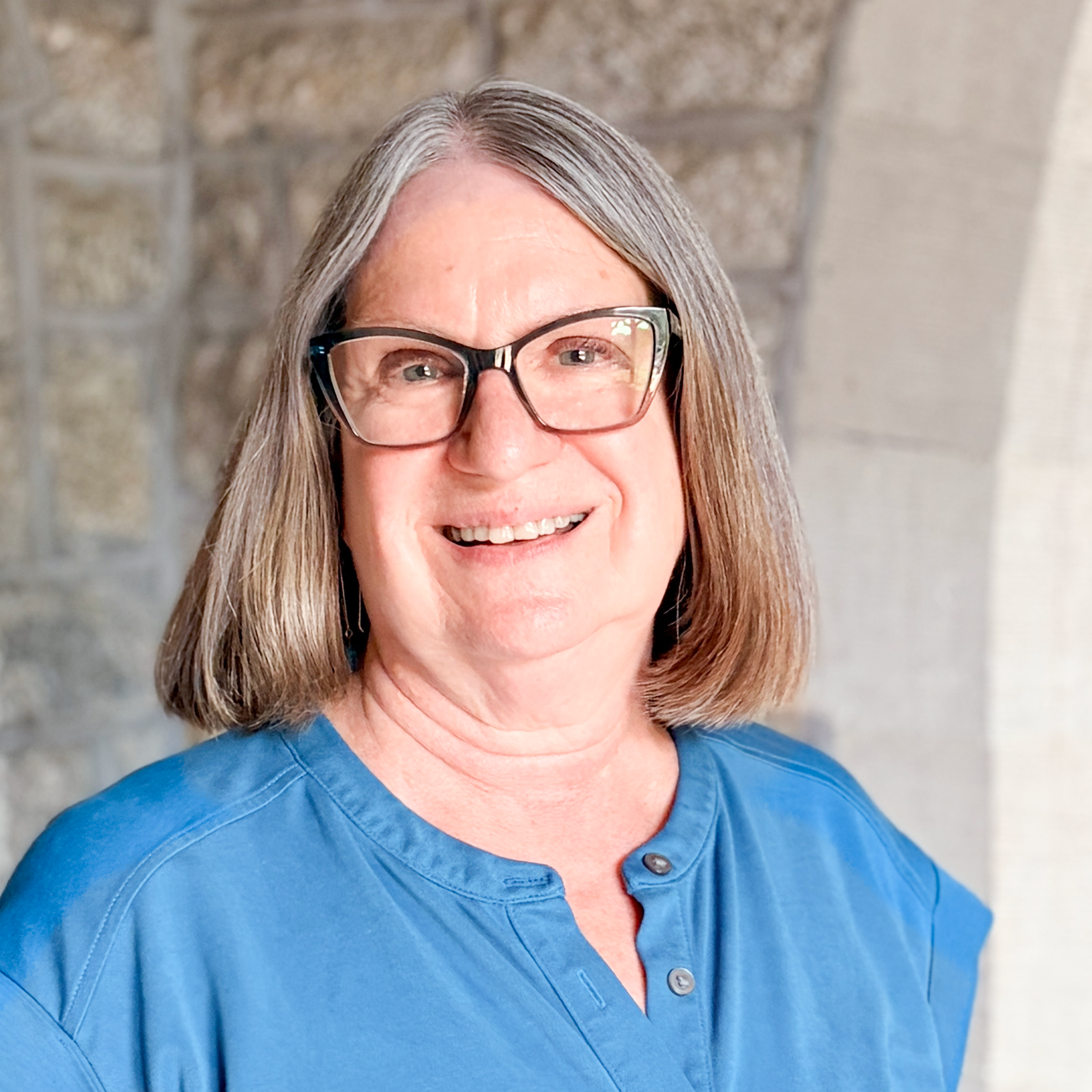 Close-up of a woman with gray hair wearing black-rimmed glasses and a blue shirt, smiling in front of a brick wall background.