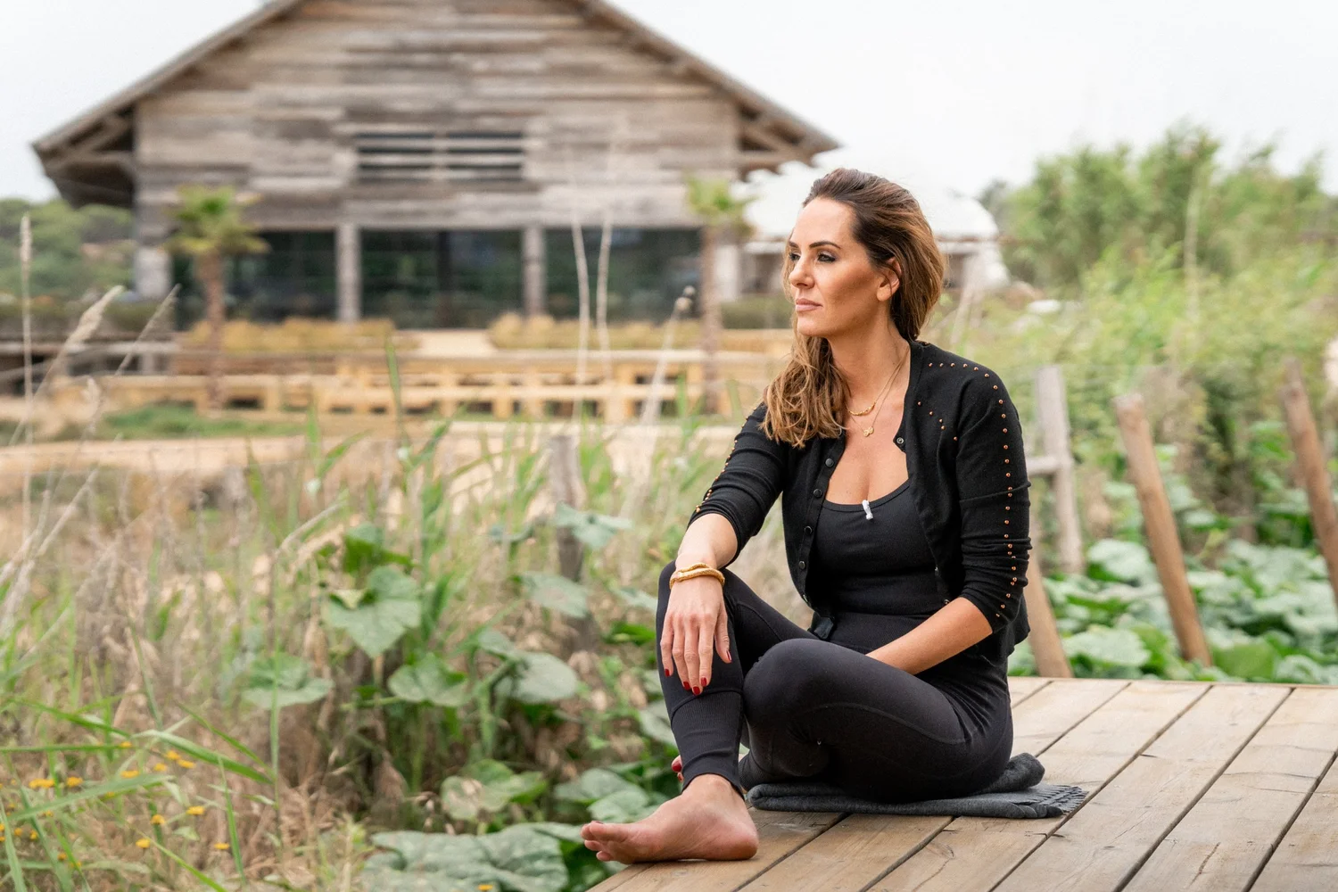 A woman sitting cross-legged on a mat on a boardwalk beside the vegetable beds, the timber restaurant building behind her.